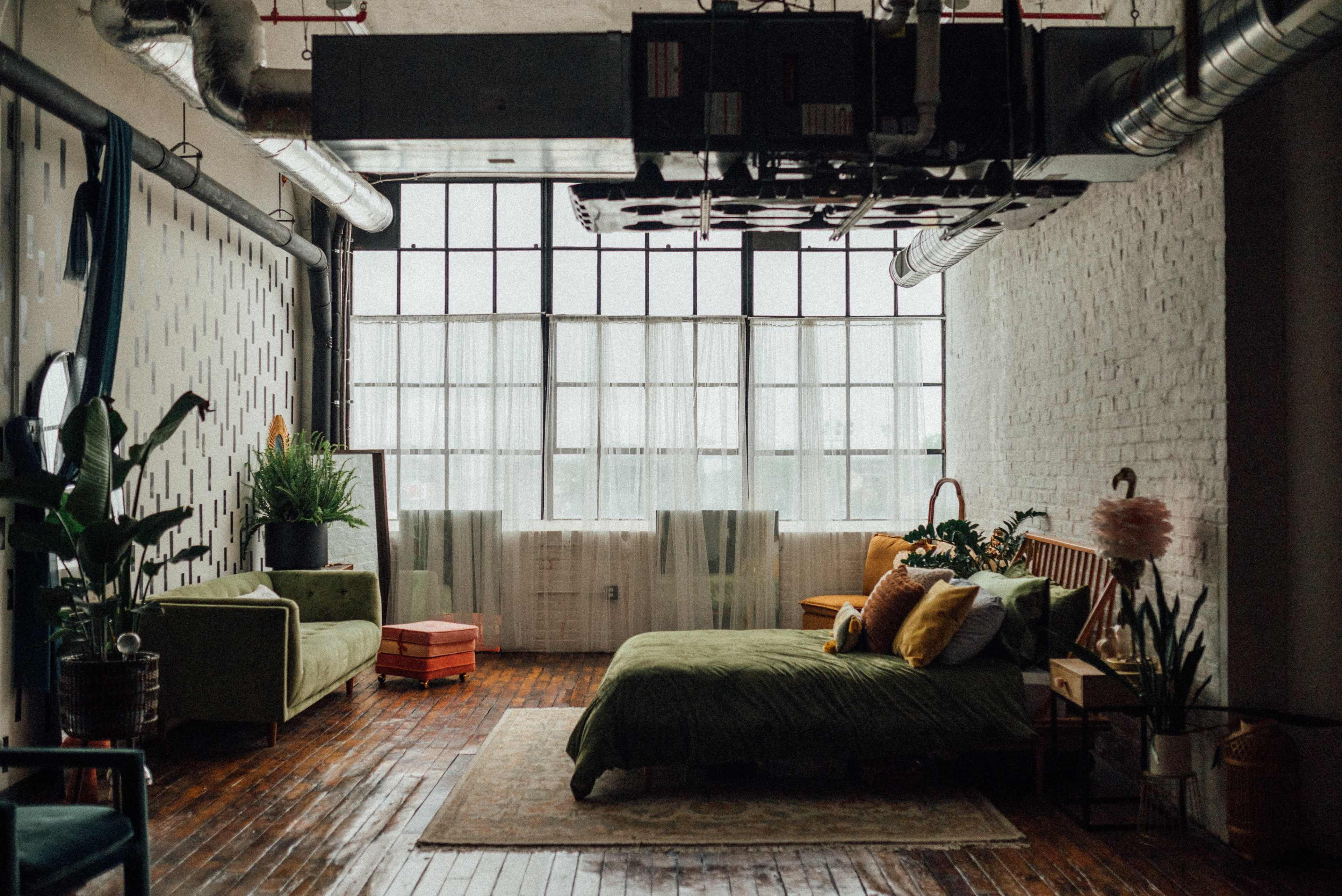 A well-lit loft bedroom features a green bedspread, wooden floors, potted plants, and large windows with sheer curtains.