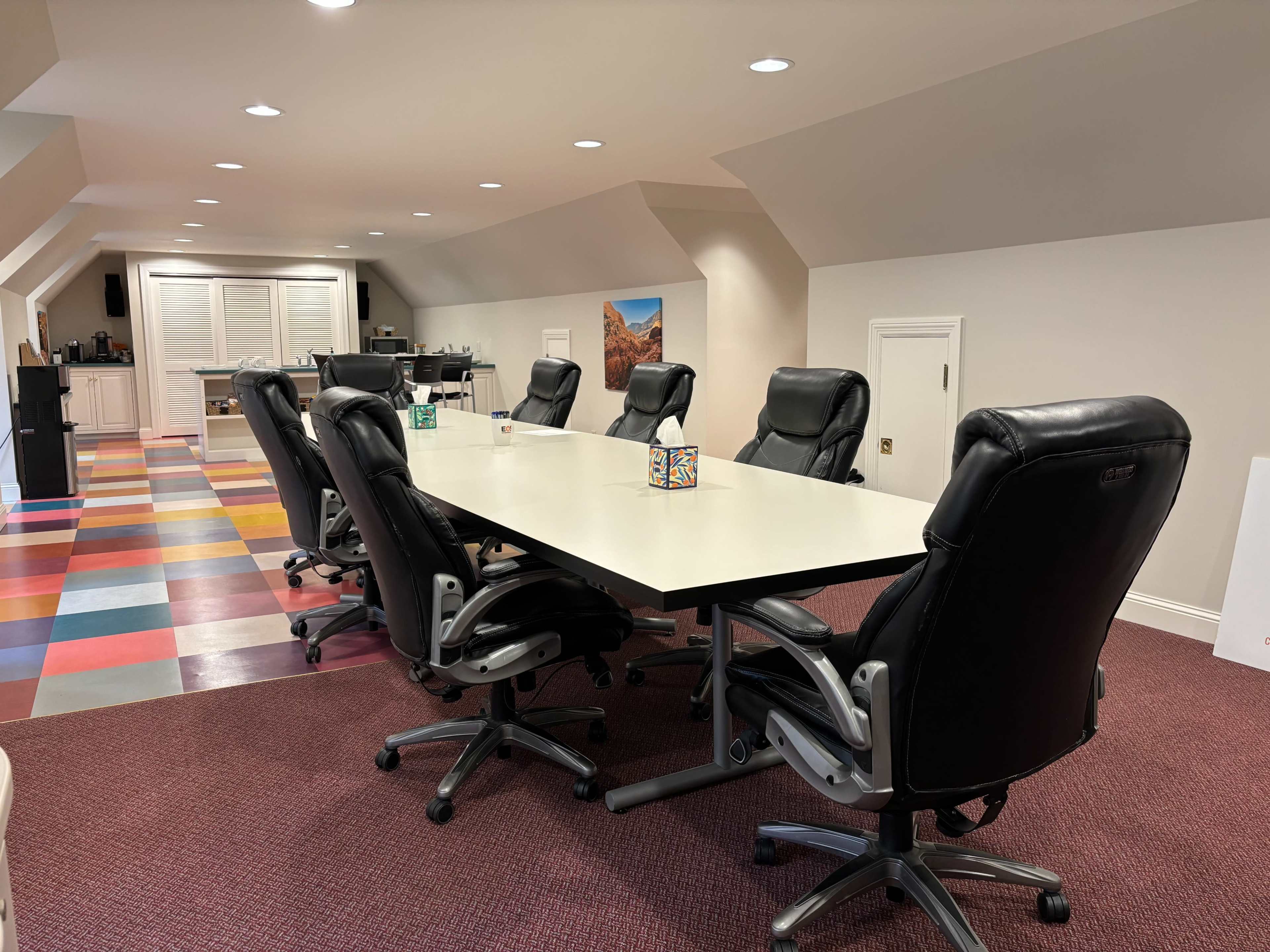 A large conference table is surrounded by black office chairs in a brightly colored meeting room with a kitchenette in the background.