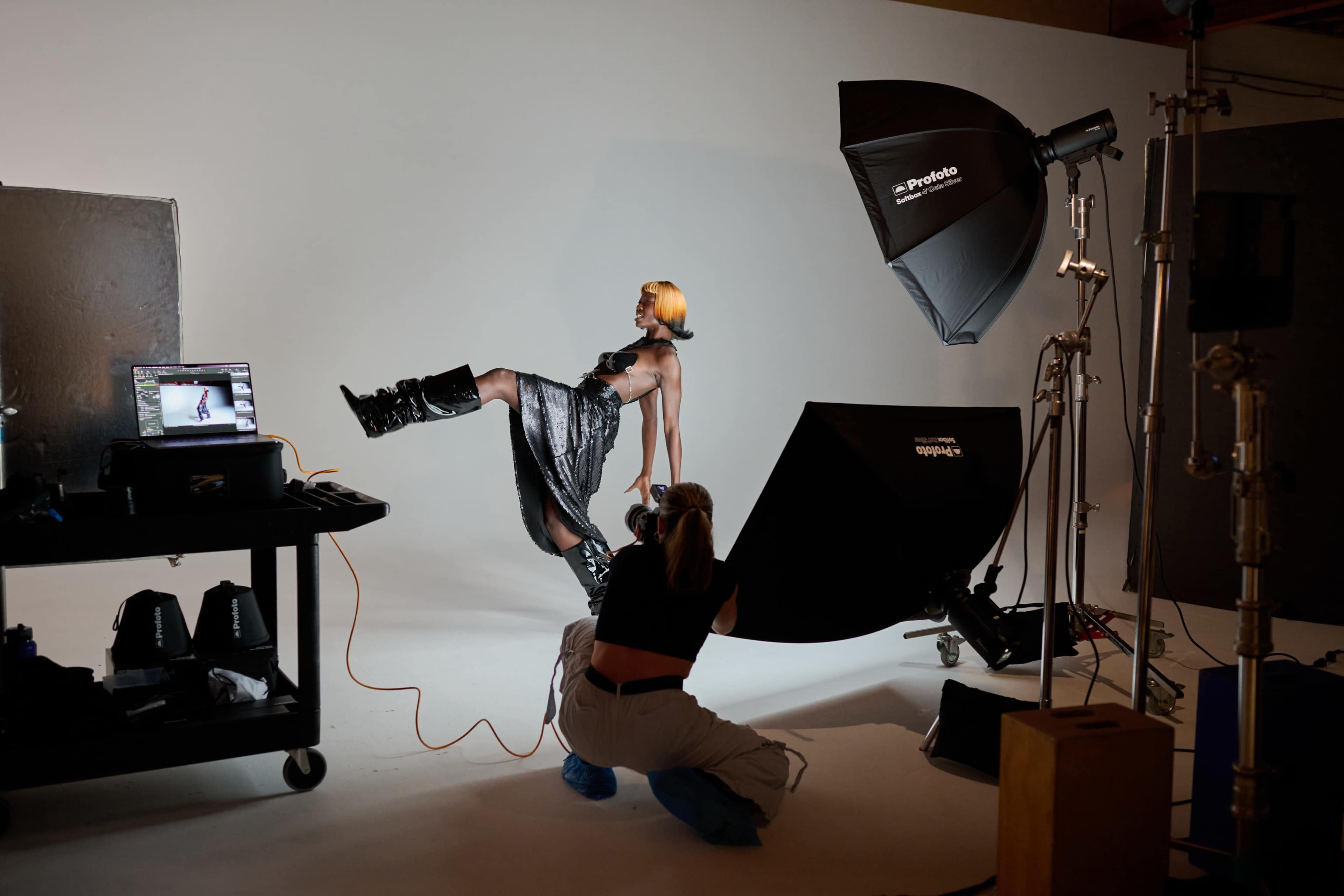 A model poses dramatically in front of a white backdrop while a photographer adjusts lighting equipment and settings.