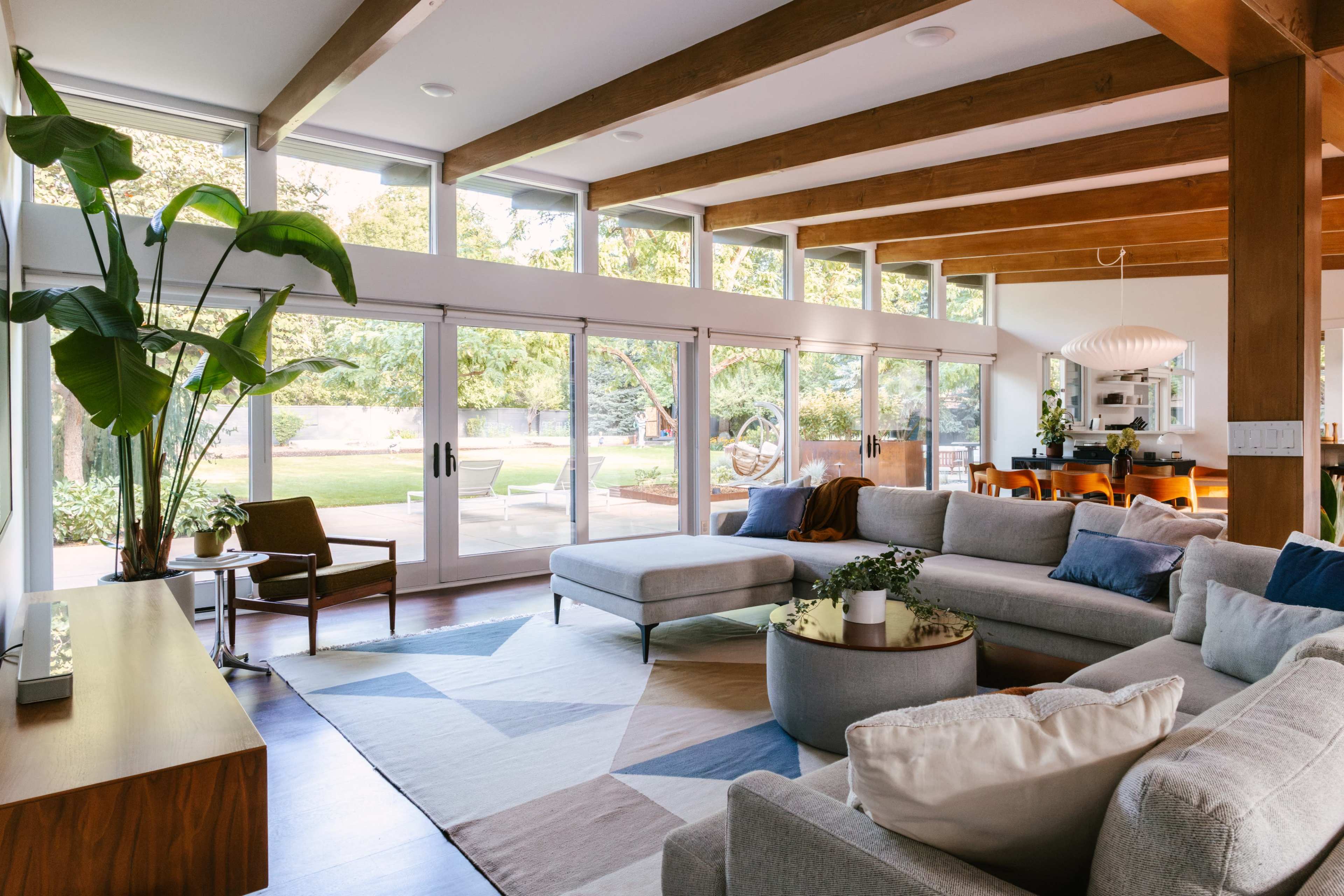 The image shows a modern living room with large windows, wooden beams, and a sectional sofa arranged around a central coffee table.