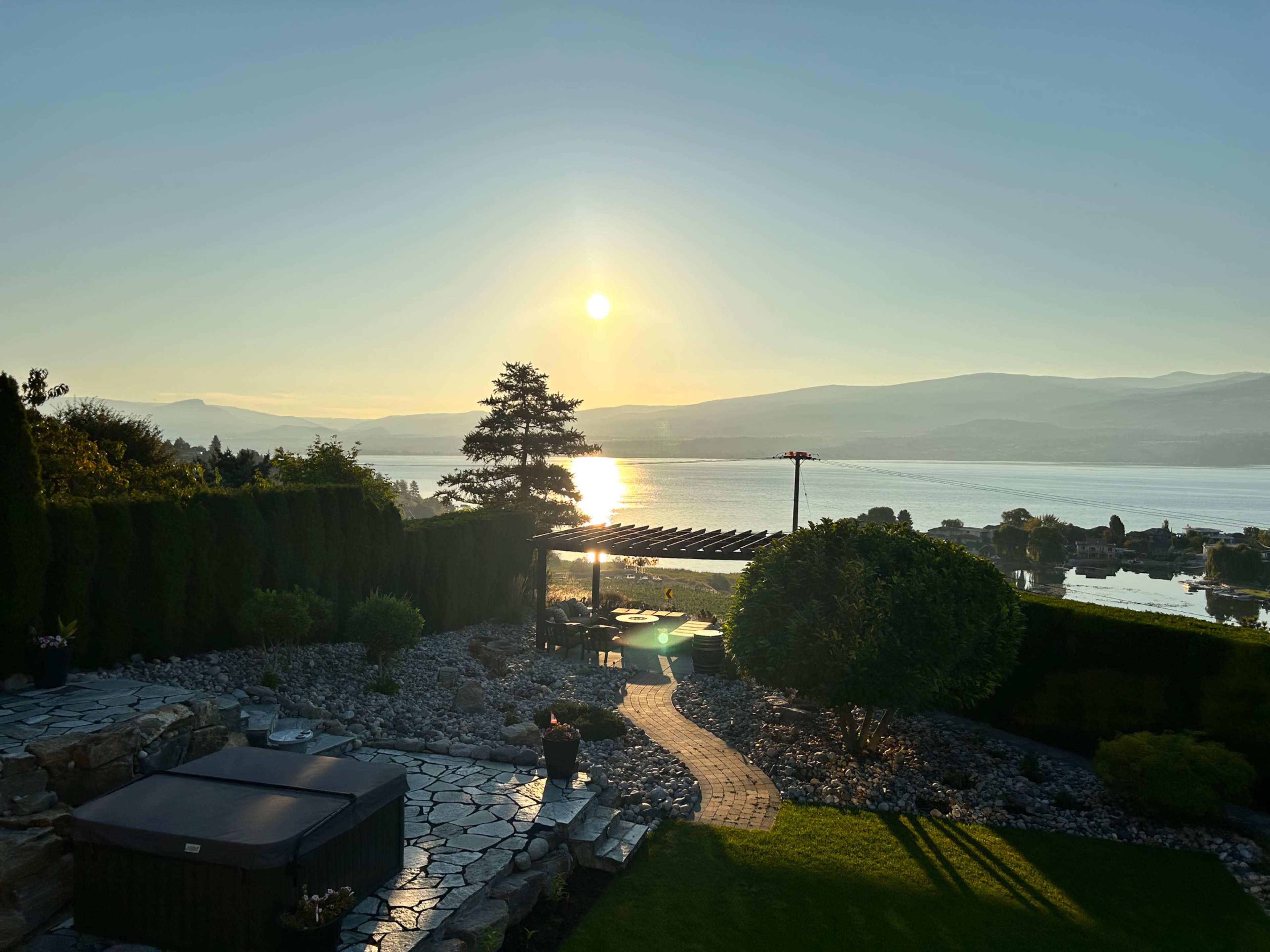 A landscaped garden with a hot tub, leading to a view of a calm lake and mountains at sunset.