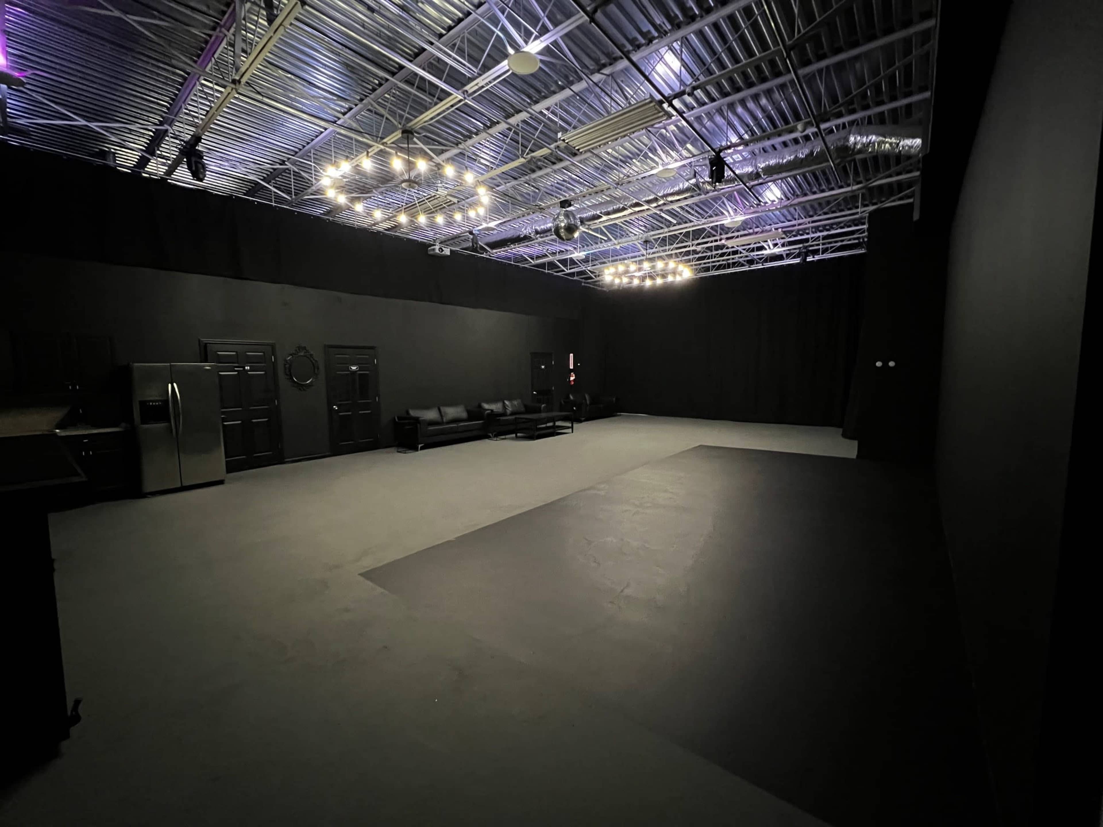 A spacious, dimly lit room with a black ceiling and walls, featuring a few lights, a sofa, and kitchen appliances in the background.