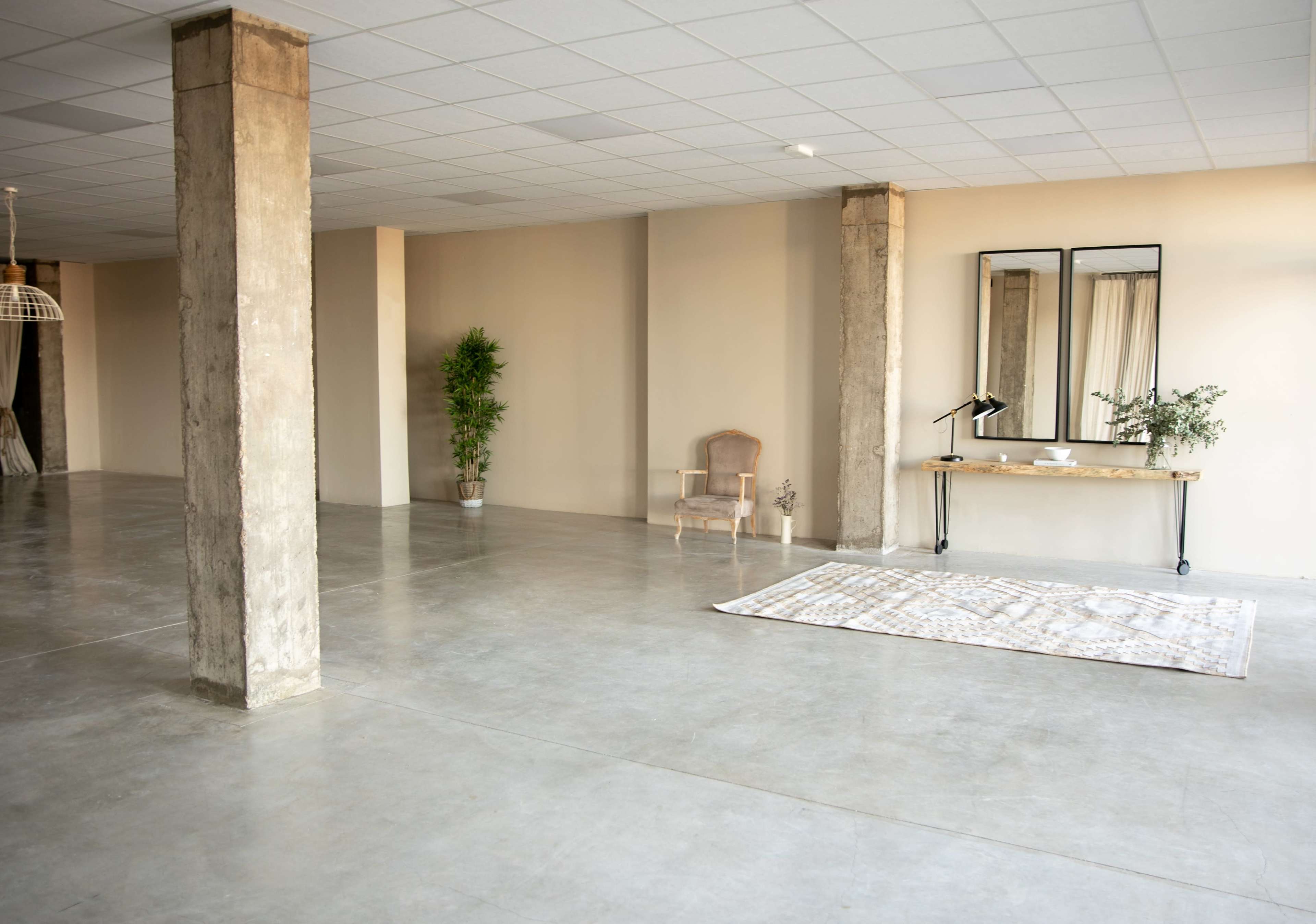 The image shows a spacious, empty interior with concrete floors, tall pillars, a small table with a lamp, a mirror, and a decorative rug.