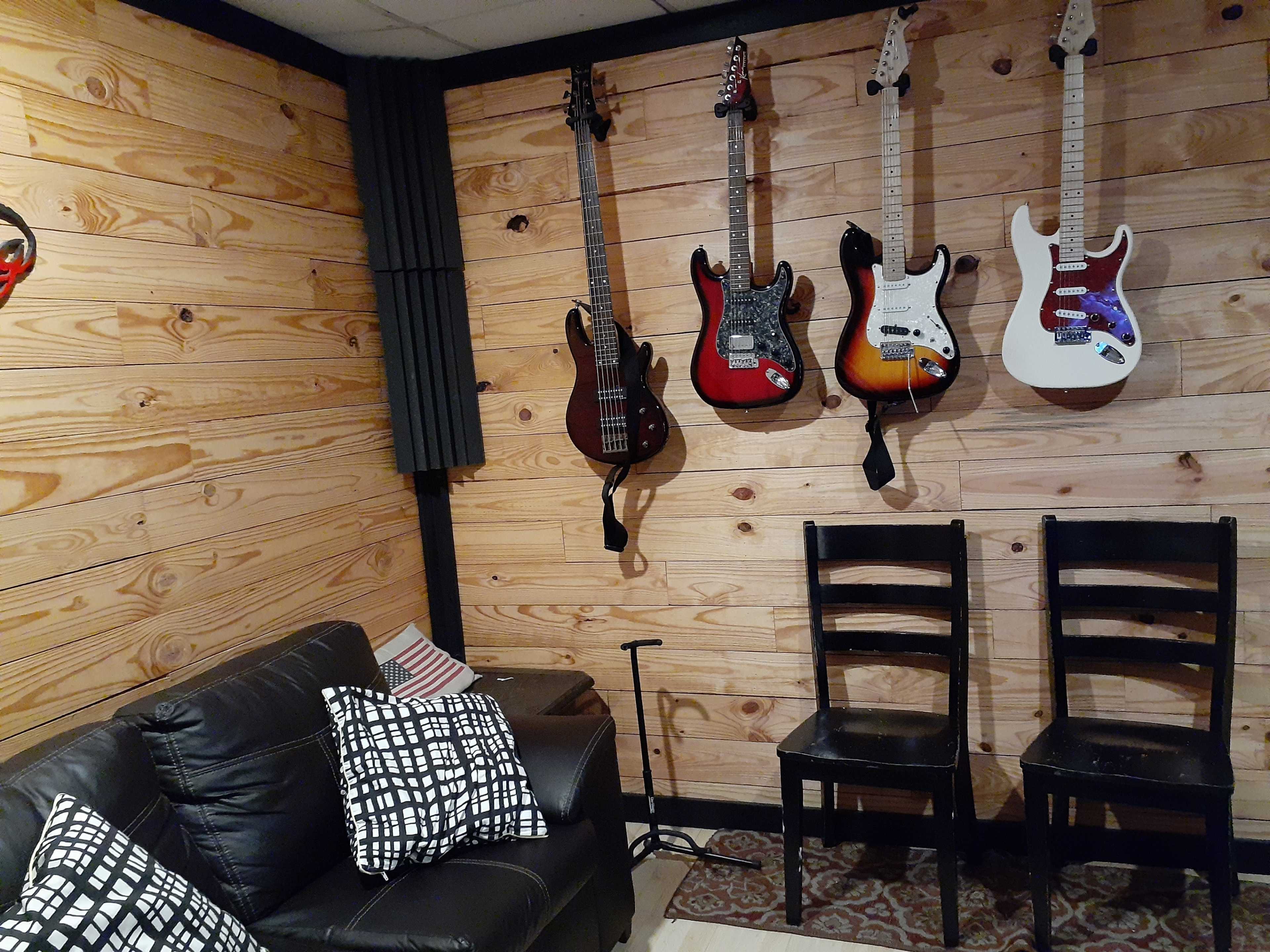A cozy music room with four guitars hanging on a wooden wall and two black chairs positioned near a dark couch.