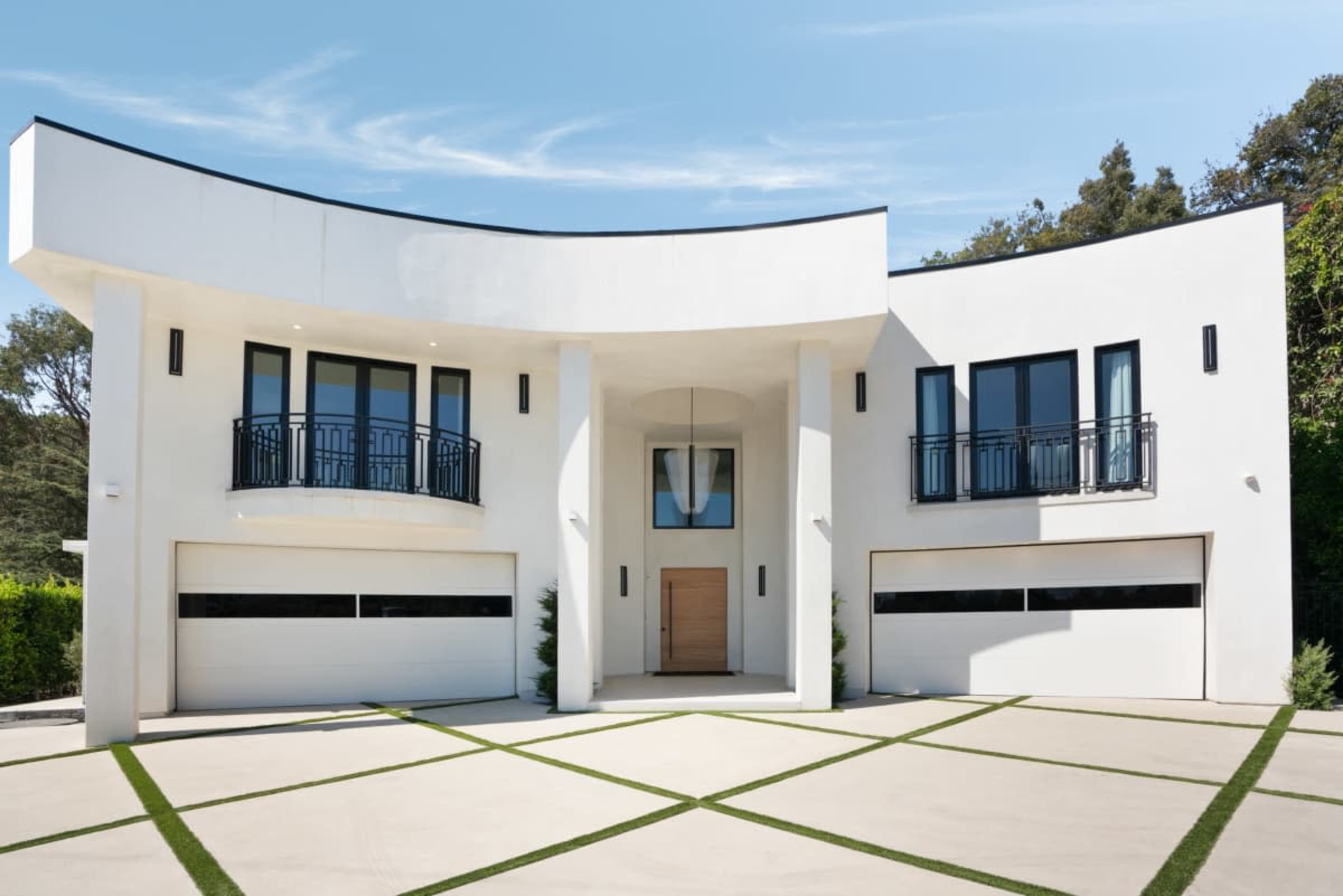The image shows a modern two-story house with a curved facade, large windows, and a symmetrical garage entrance.
