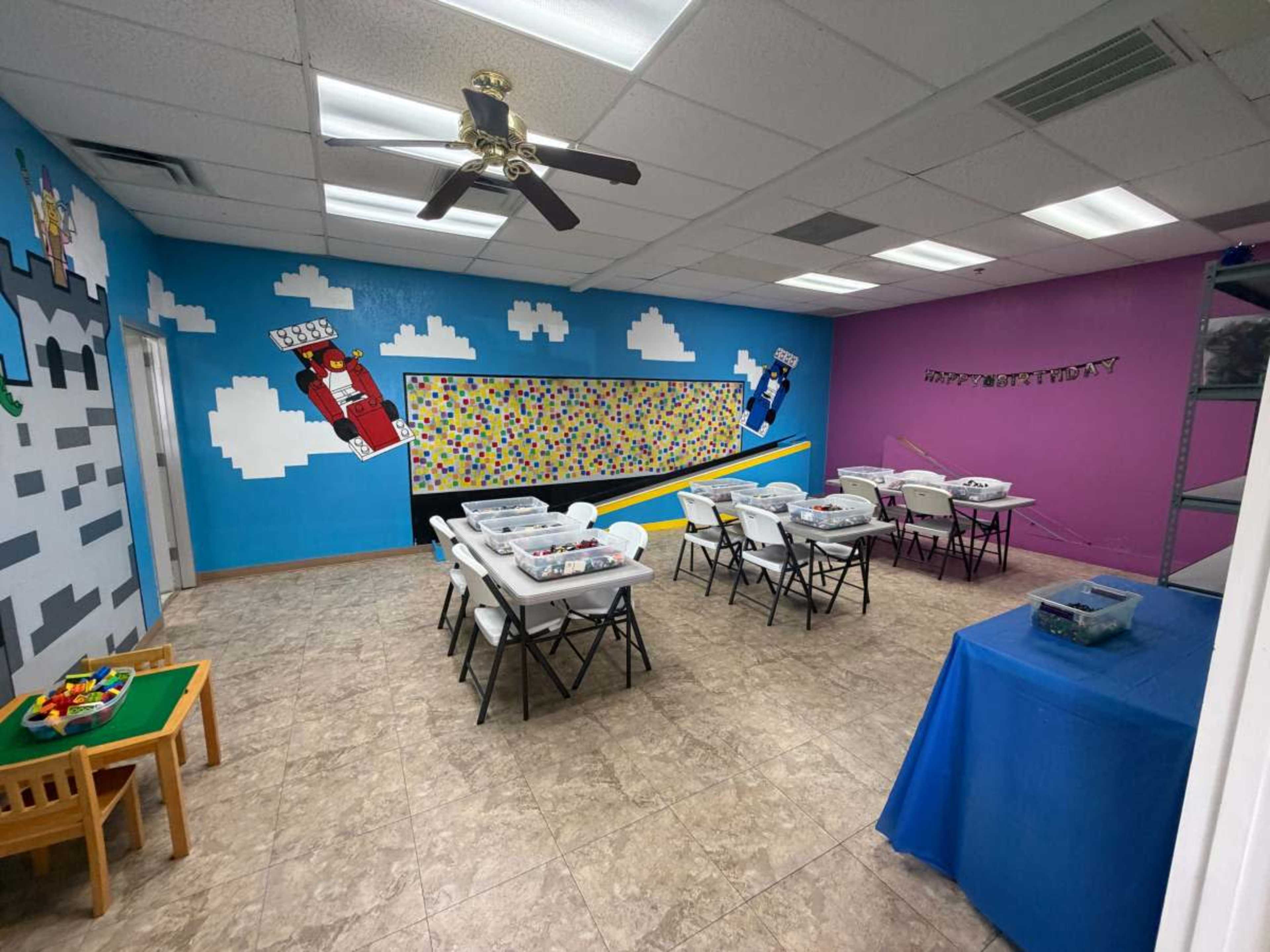 The image shows a brightly colored classroom with a large mural on one wall, several folding tables with chairs set up, and shelves along one side.