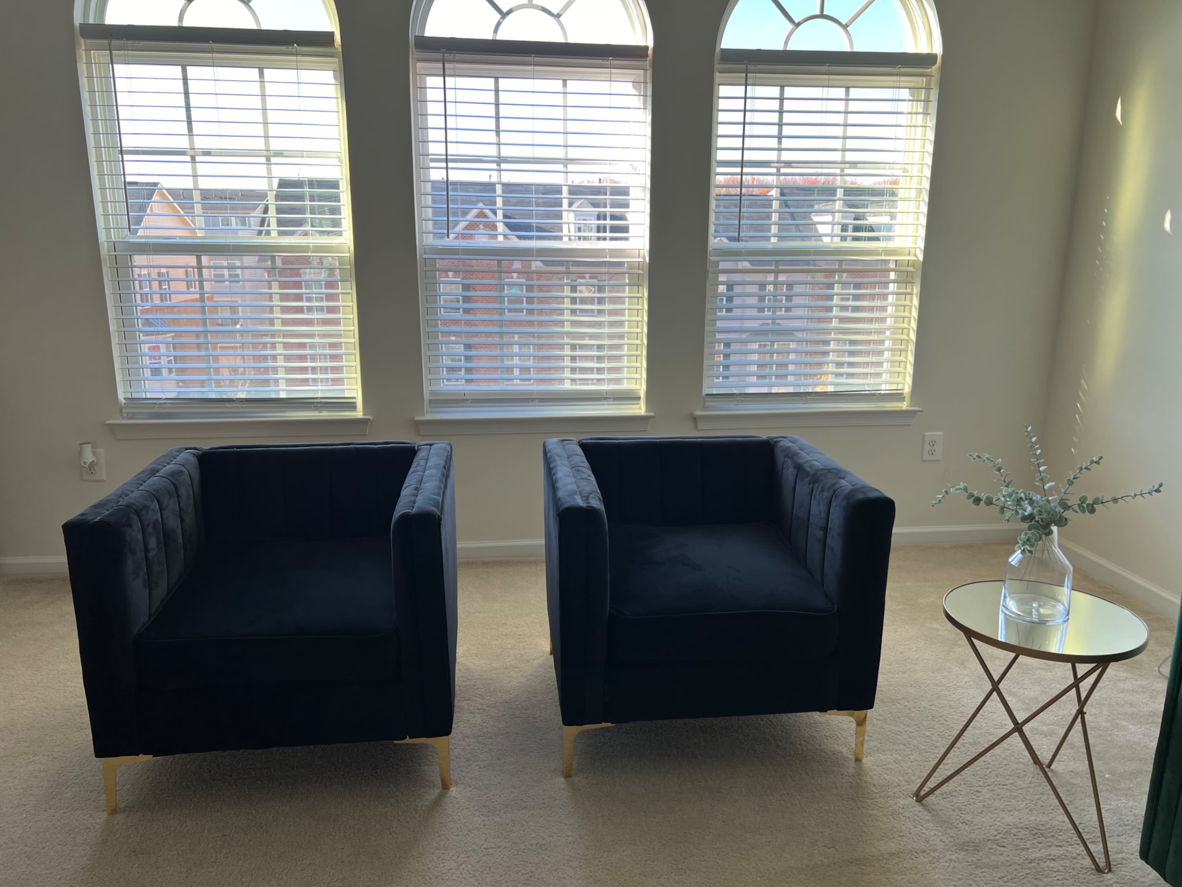 Two black armchairs are positioned opposite each other beside a small glass side table in a well-lit room with three large windows.
