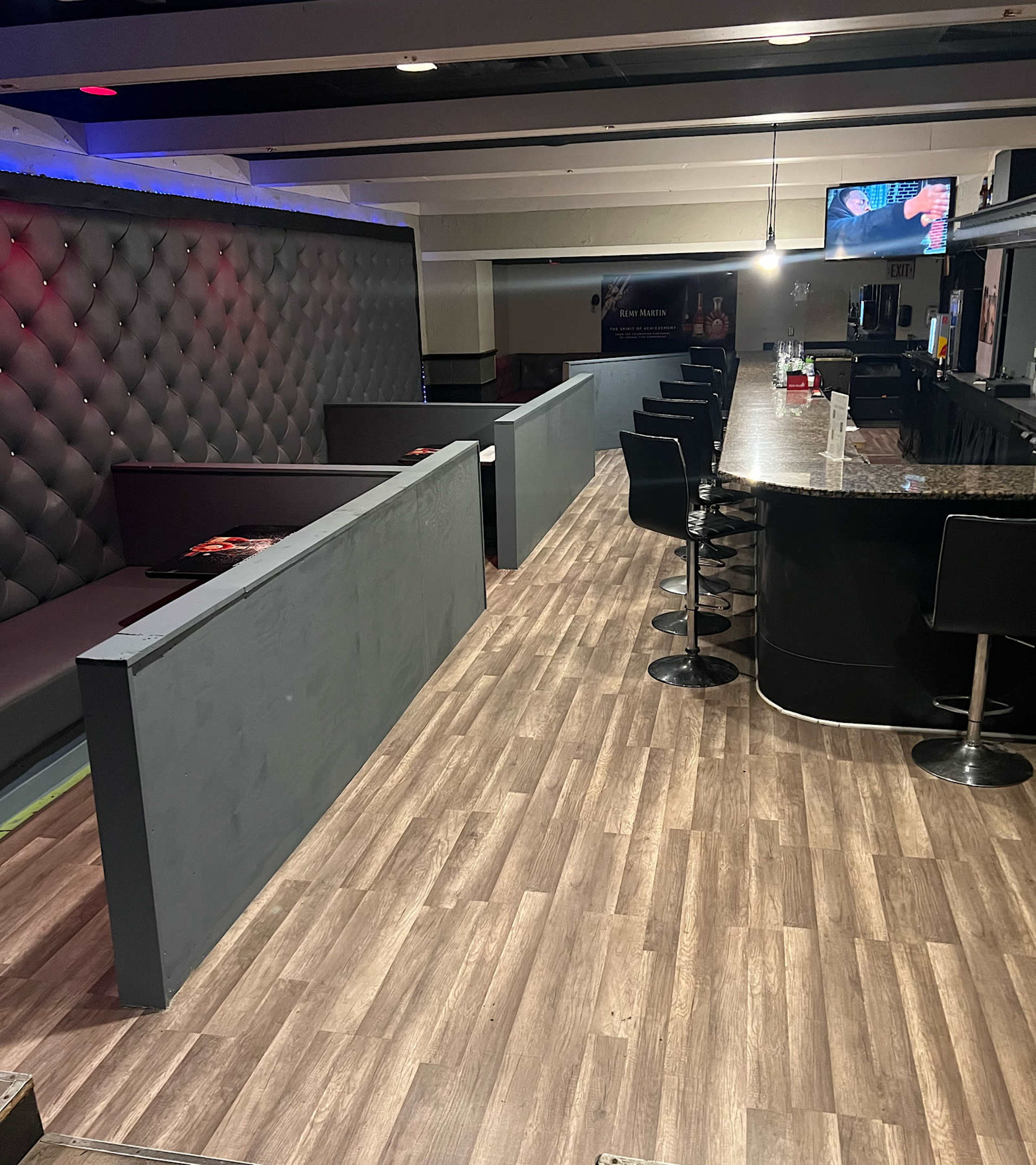 A bar area with modern seating arrangements and a polished counter, featuring a gray tufted wall and hardwood flooring.