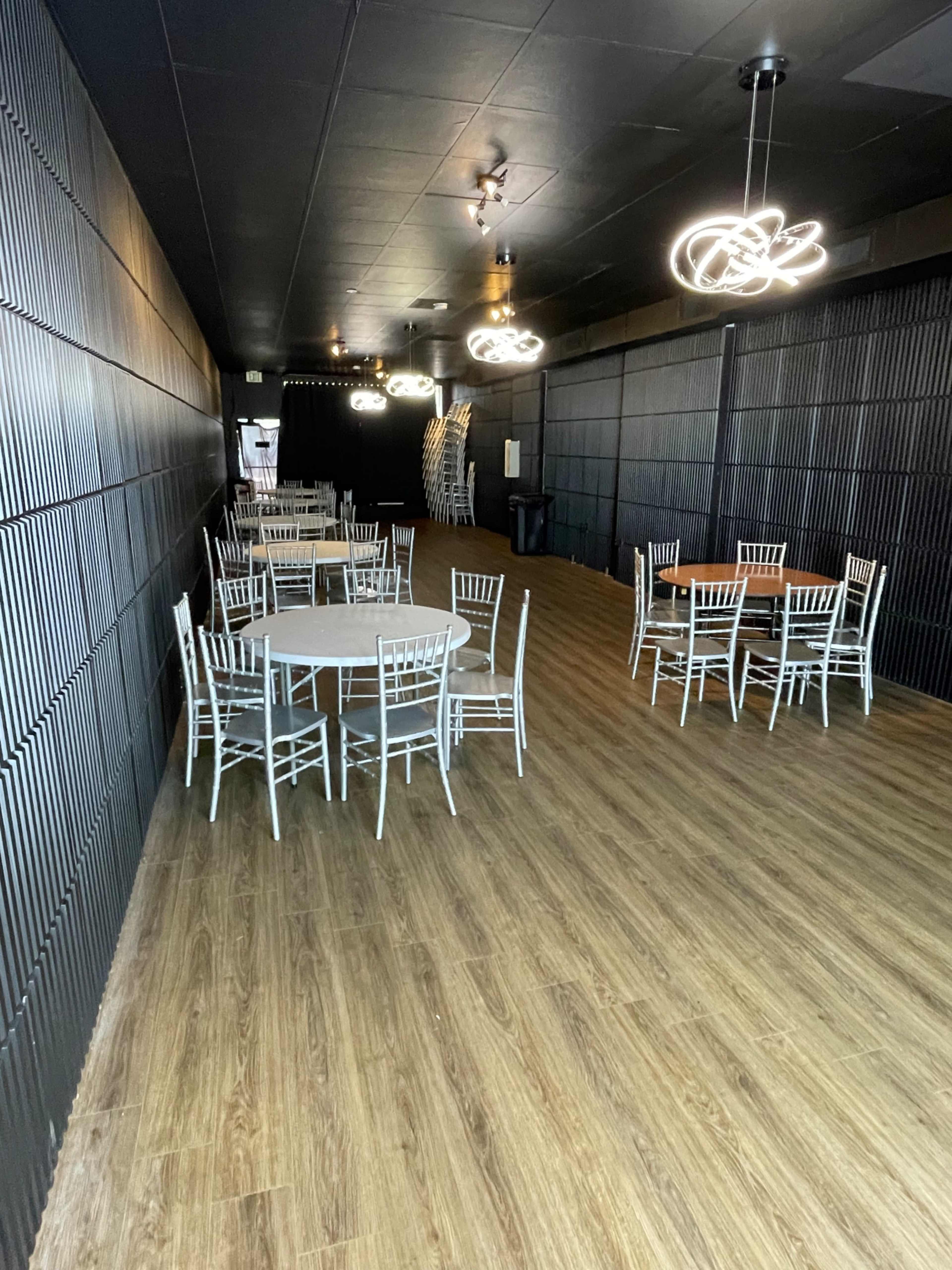 A spacious, dimly-lit event venue with several round tables surrounded by white chairs and modern light fixtures hanging from the ceiling.