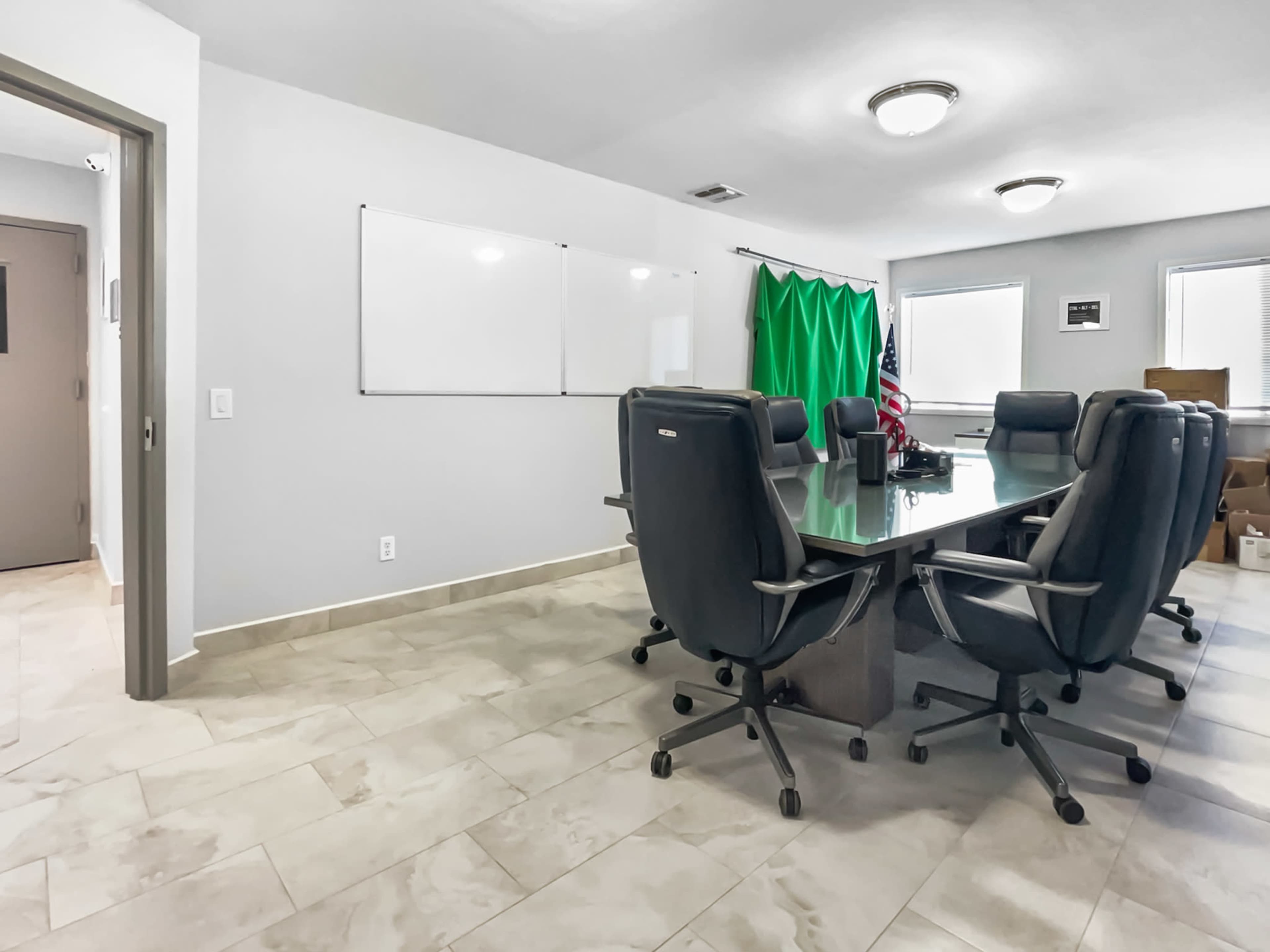 A meeting room features a large glass table surrounded by black office chairs, with a whiteboard and a green backdrop on the walls.