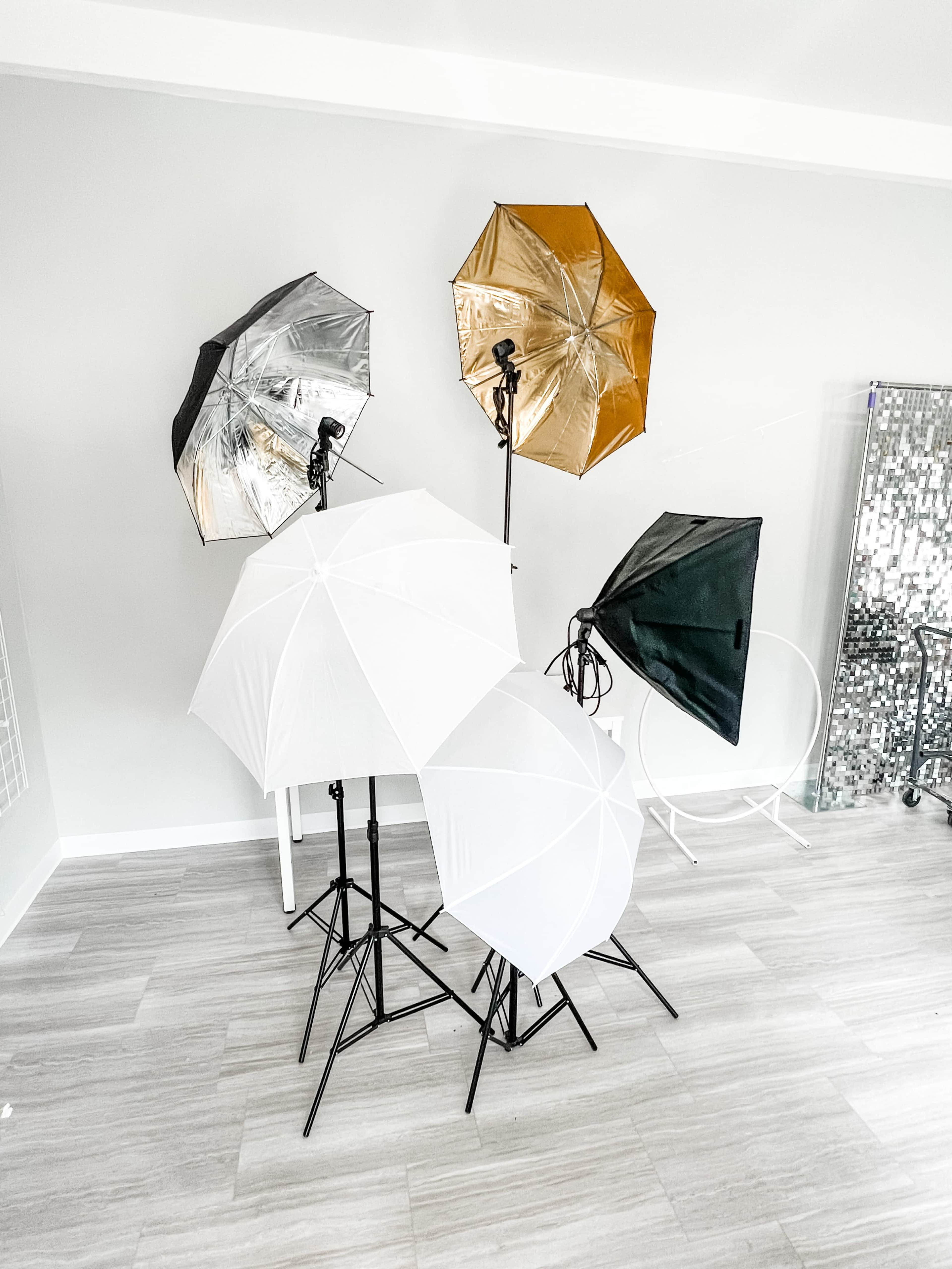 Multiple photography lighting umbrellas, including white, silver, and gold, set up on stands in a well-lit room with light-colored flooring.