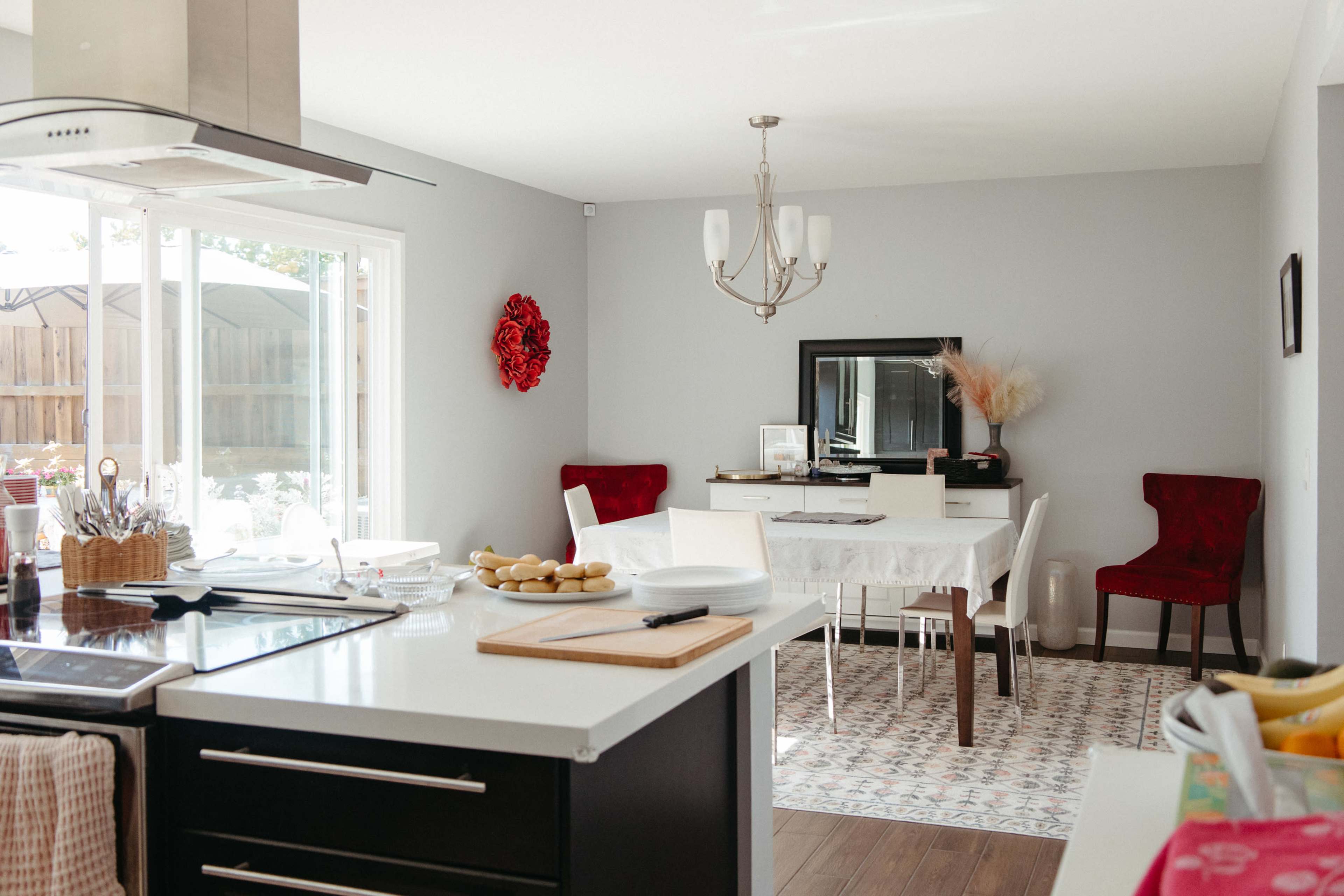 The image shows a modern kitchen and dining area with a table set for meals, red chairs, and decorative elements on the walls.