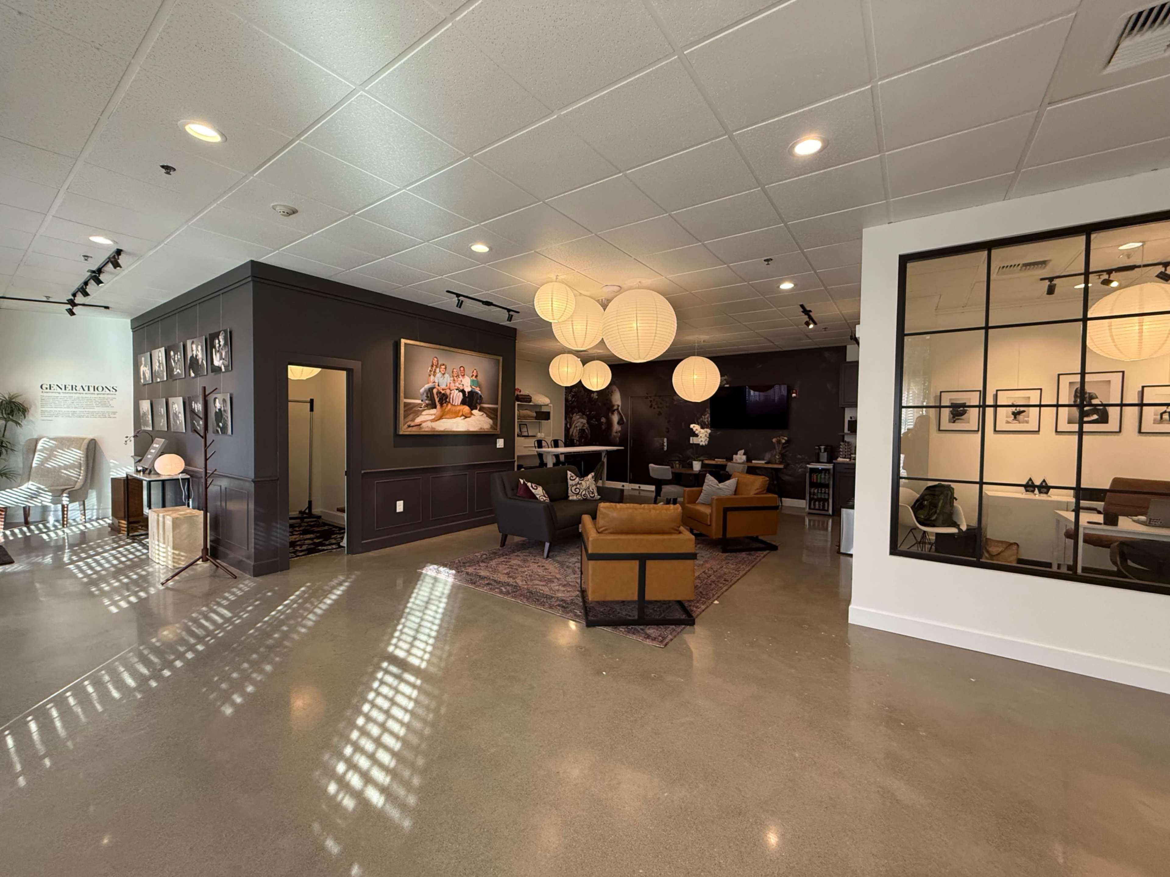 The image shows a spacious, modern interior featuring a seating area with two orange armchairs and gray sofas, framed photographs on the walls, and large pendant lights hanging from the ceiling.
