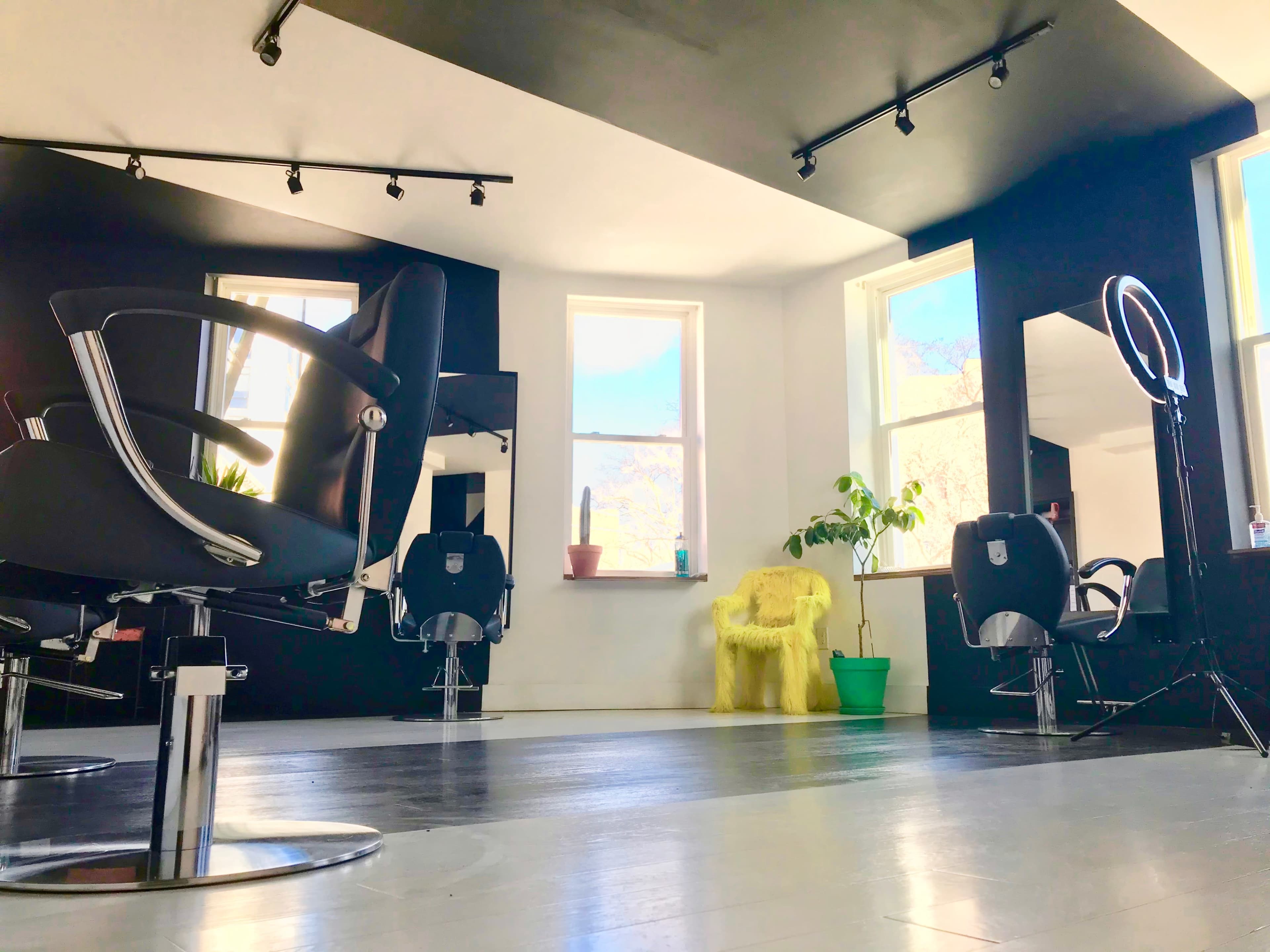 The image shows a modern hair salon with several black styling chairs, large windows, and a yellow armchair in the corner.