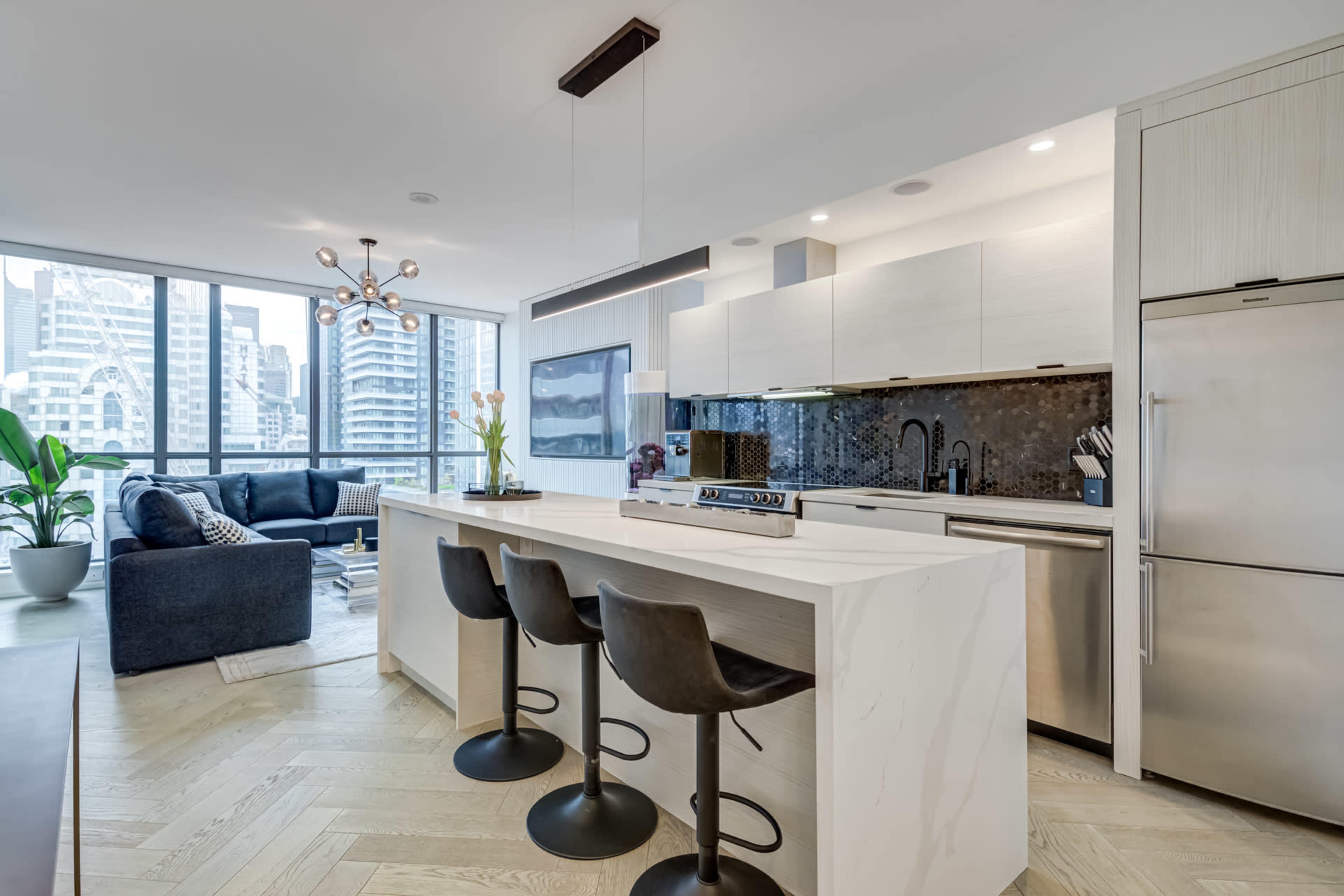 A modern kitchen features a central island with three black stools, stainless steel appliances, and large windows showcasing a city skyline.