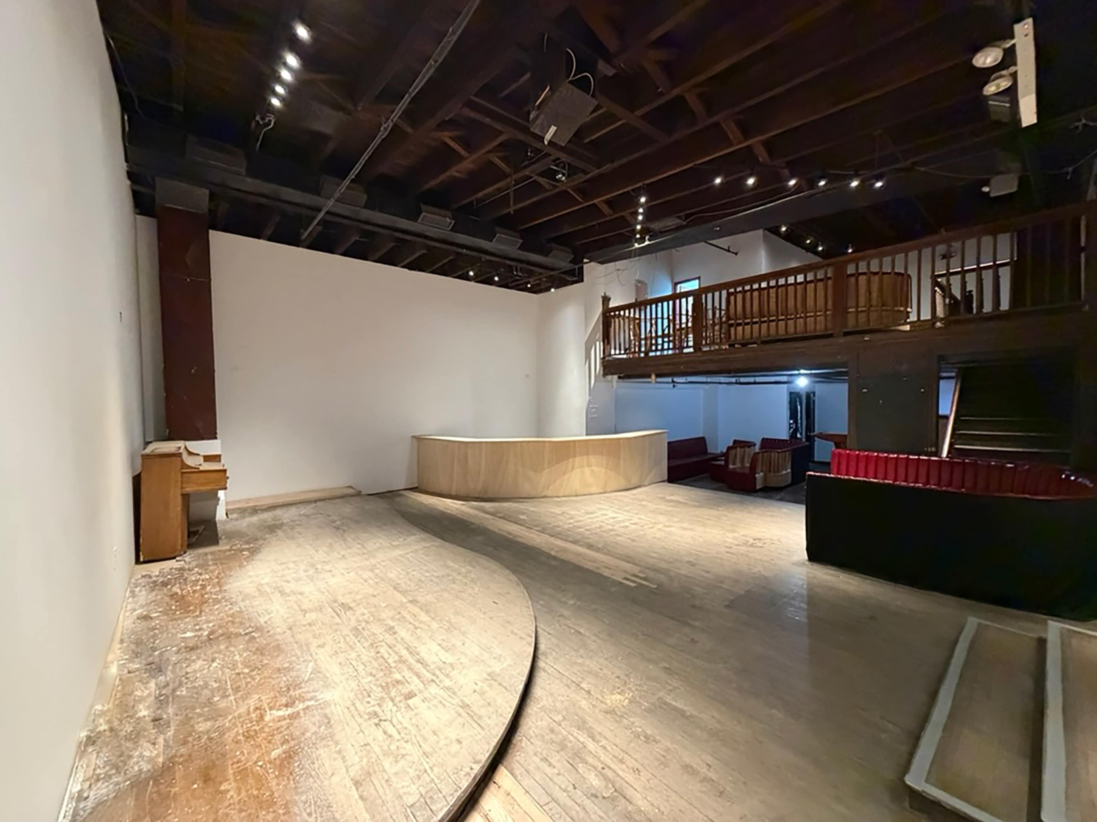 The image shows an empty auditorium with a wooden stage, a raised balcony, and sparse seating on the lower level.
