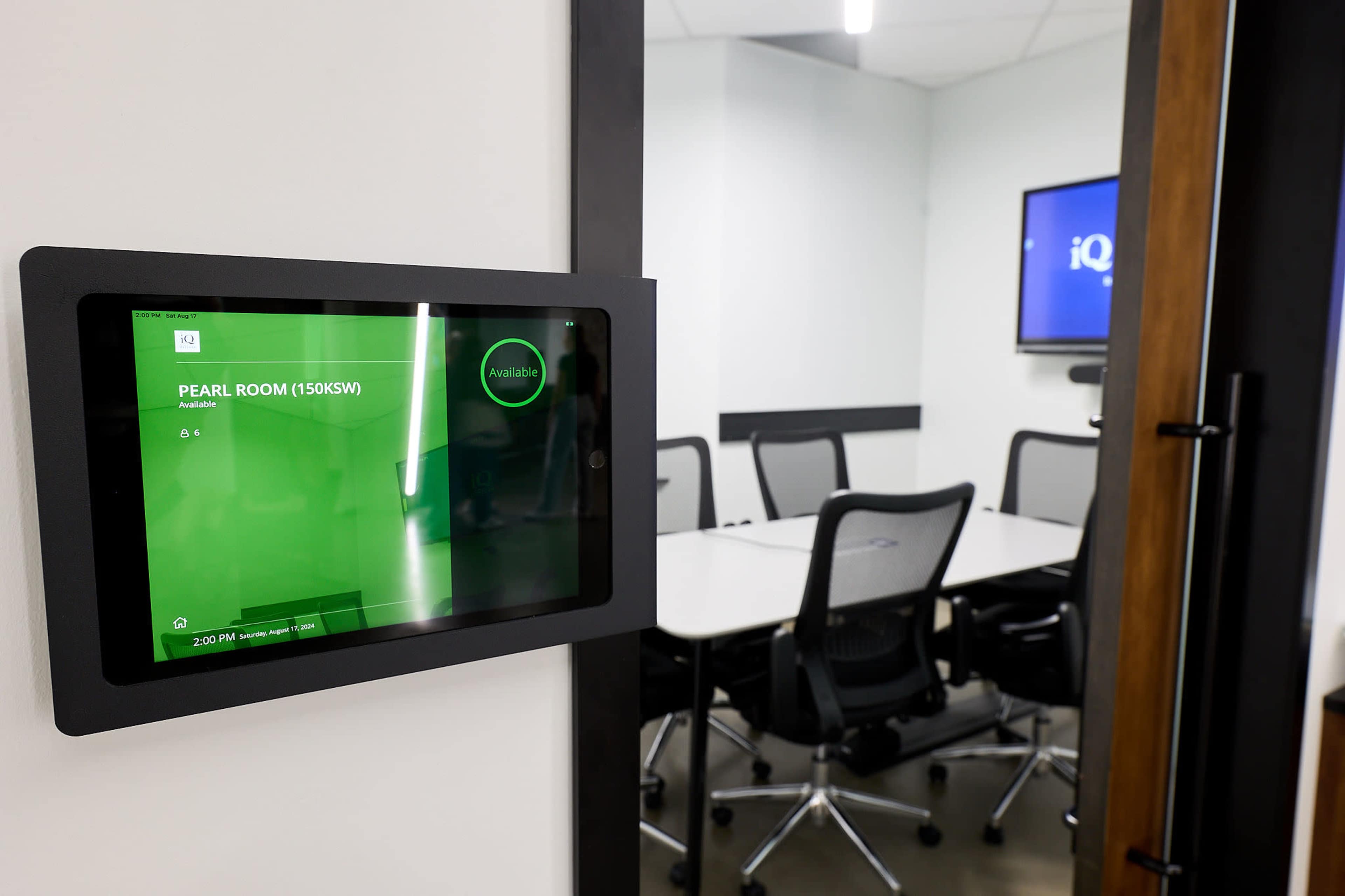 A digital room booking display is mounted on a wall next to a conference room door, showing the availability status of the "Pearl Room."
