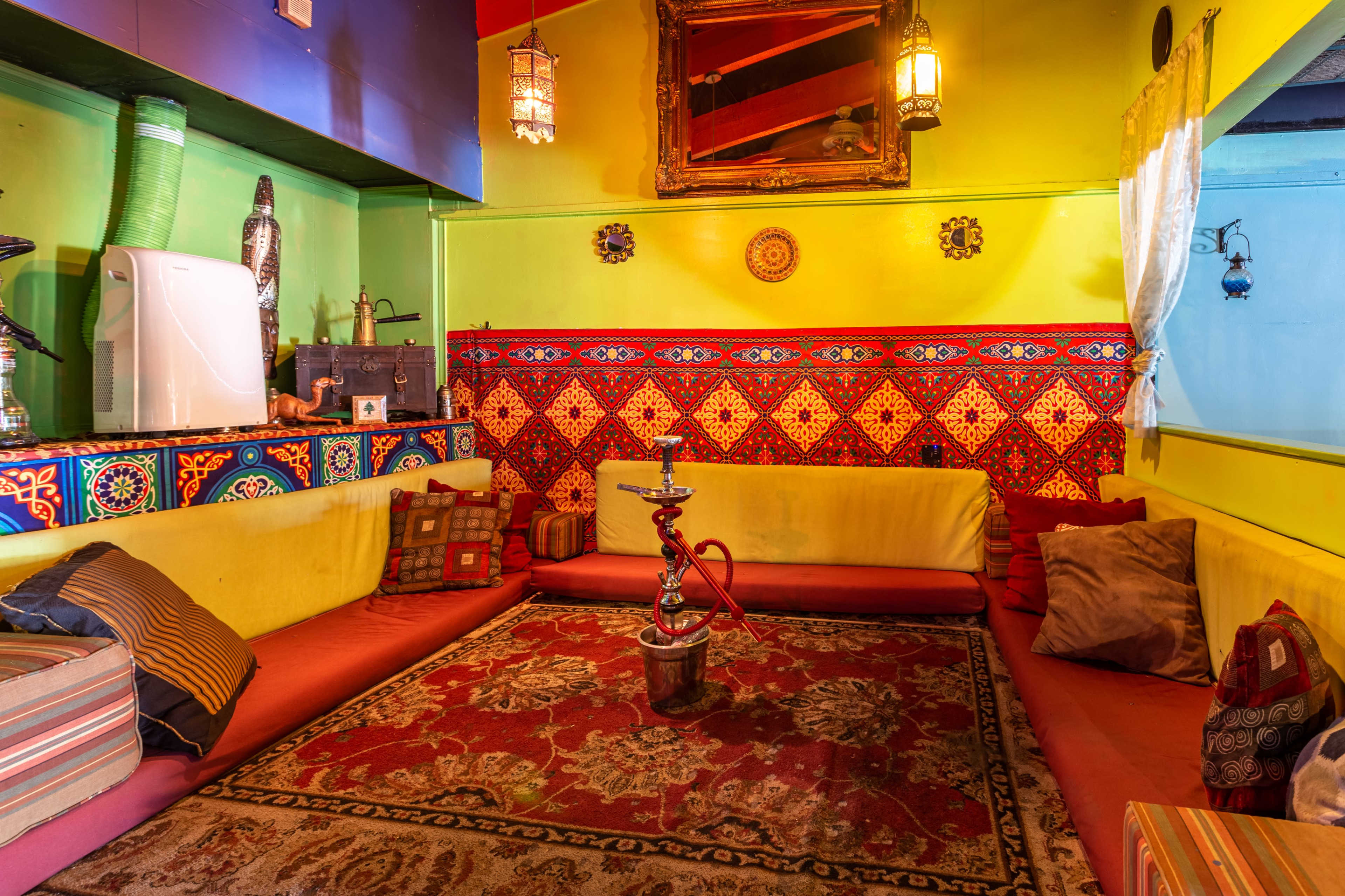 A vibrant lounge area with colorful walls, a traditional rug on the floor, low seating with cushions, and a hookah on a small table.