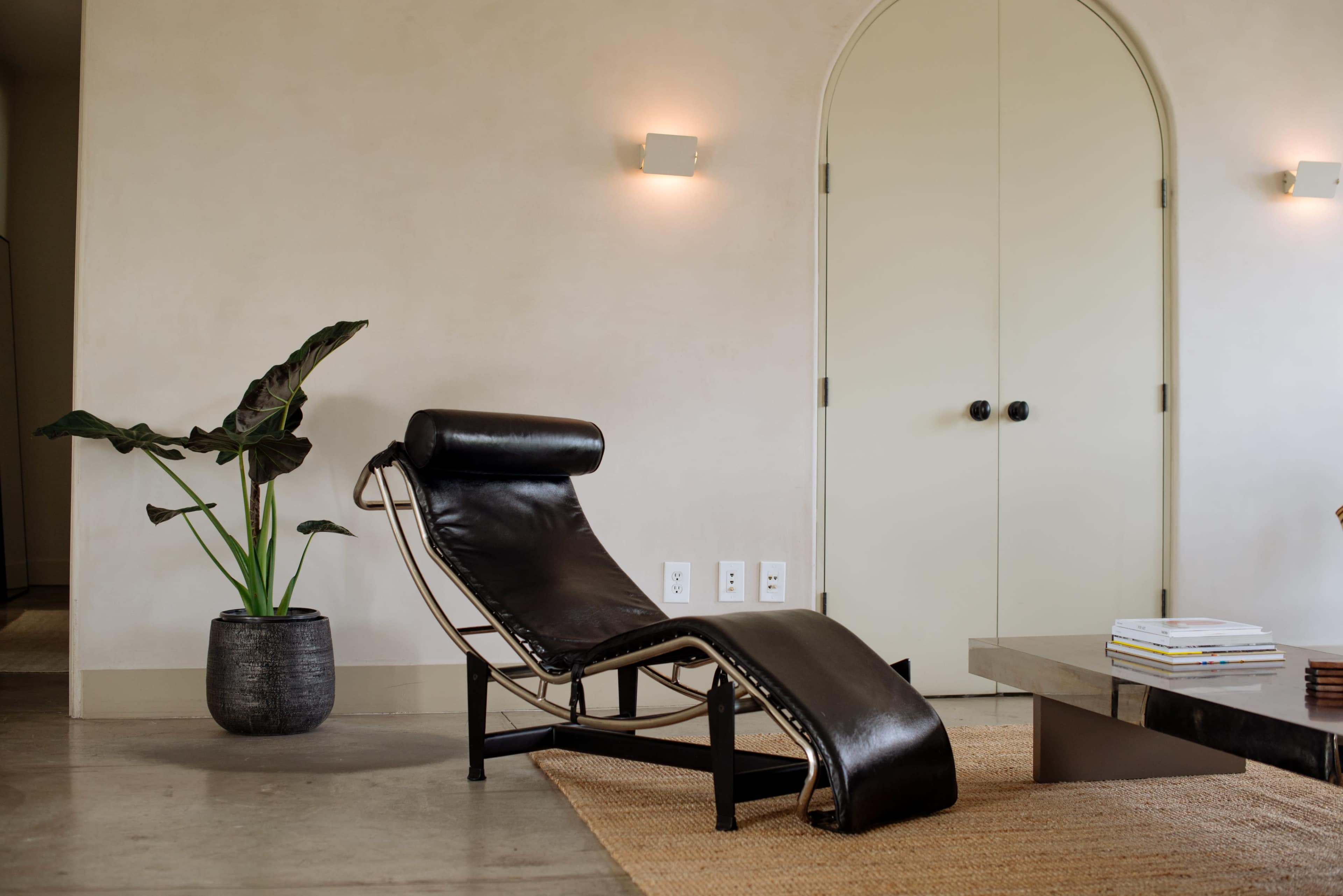 A sleek black chaise lounge sits next to a potted plant in a minimalistic room with a light-colored wall and an arched doorway.
