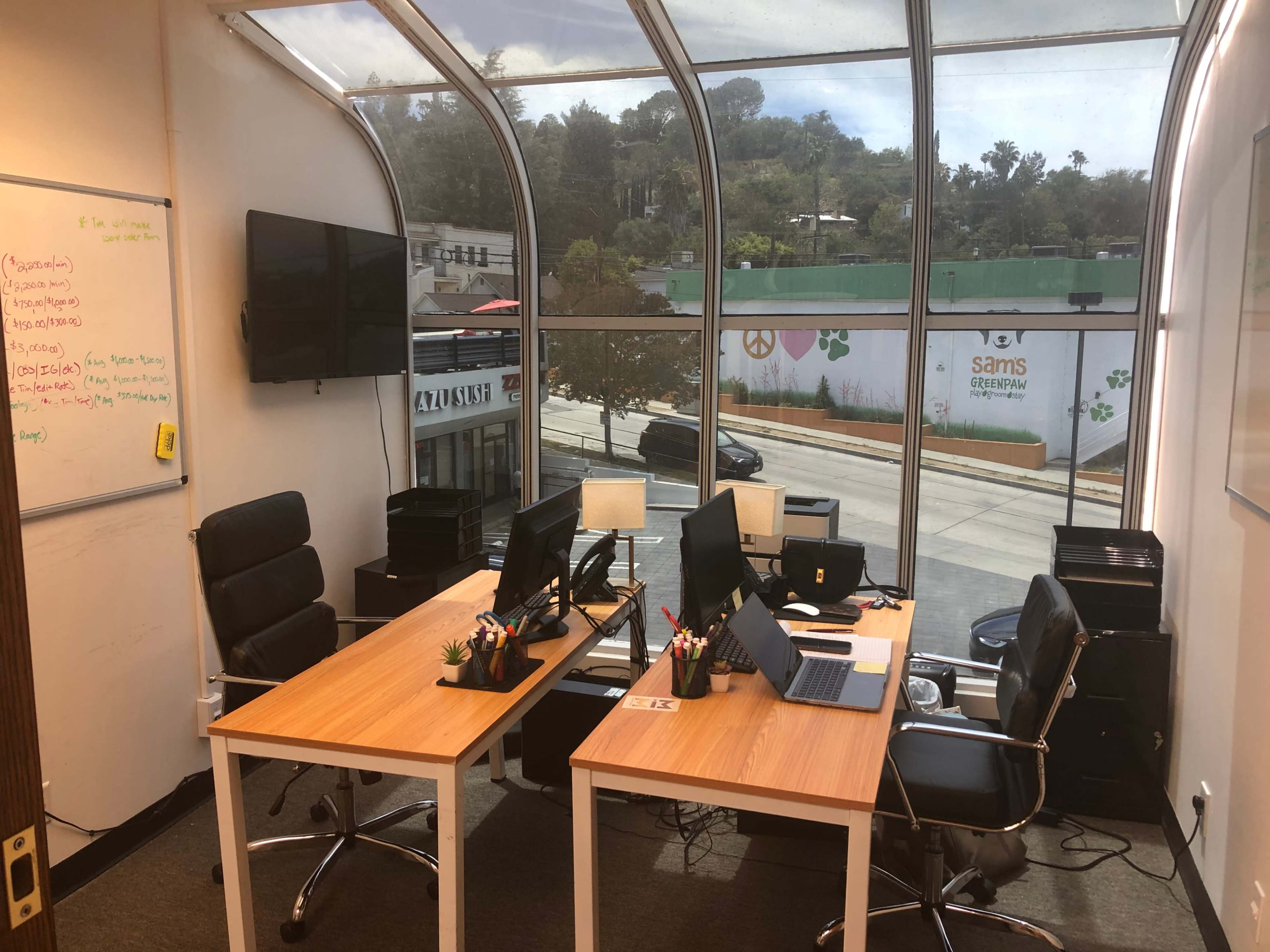 A modern office workspace with two desks, each equipped with computers, facing a large window that overlooks a street and greenery outside.