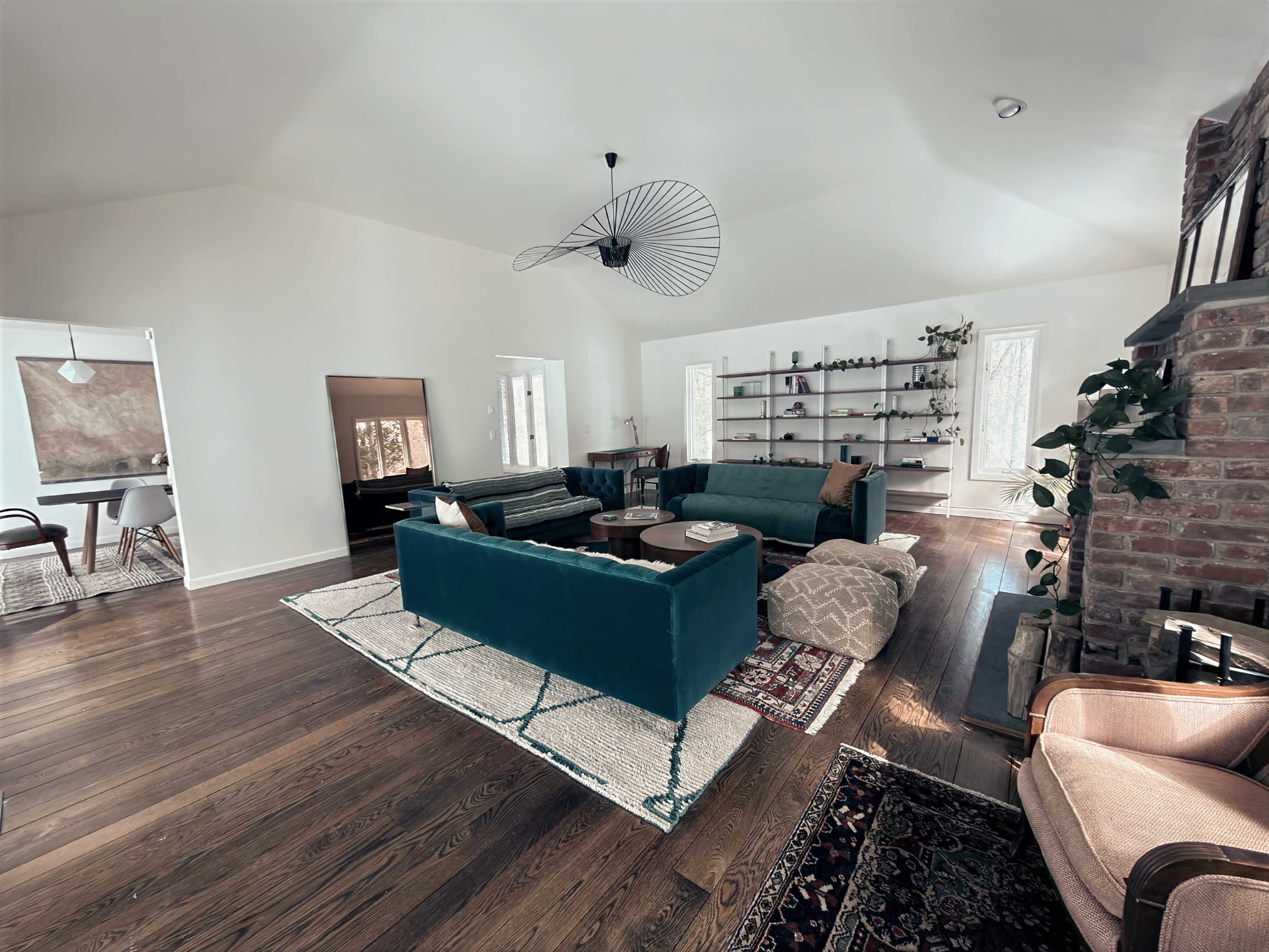 A modern living room features teal sofas, a round coffee table, a bookshelf with decorative items, and large windows allowing natural light.