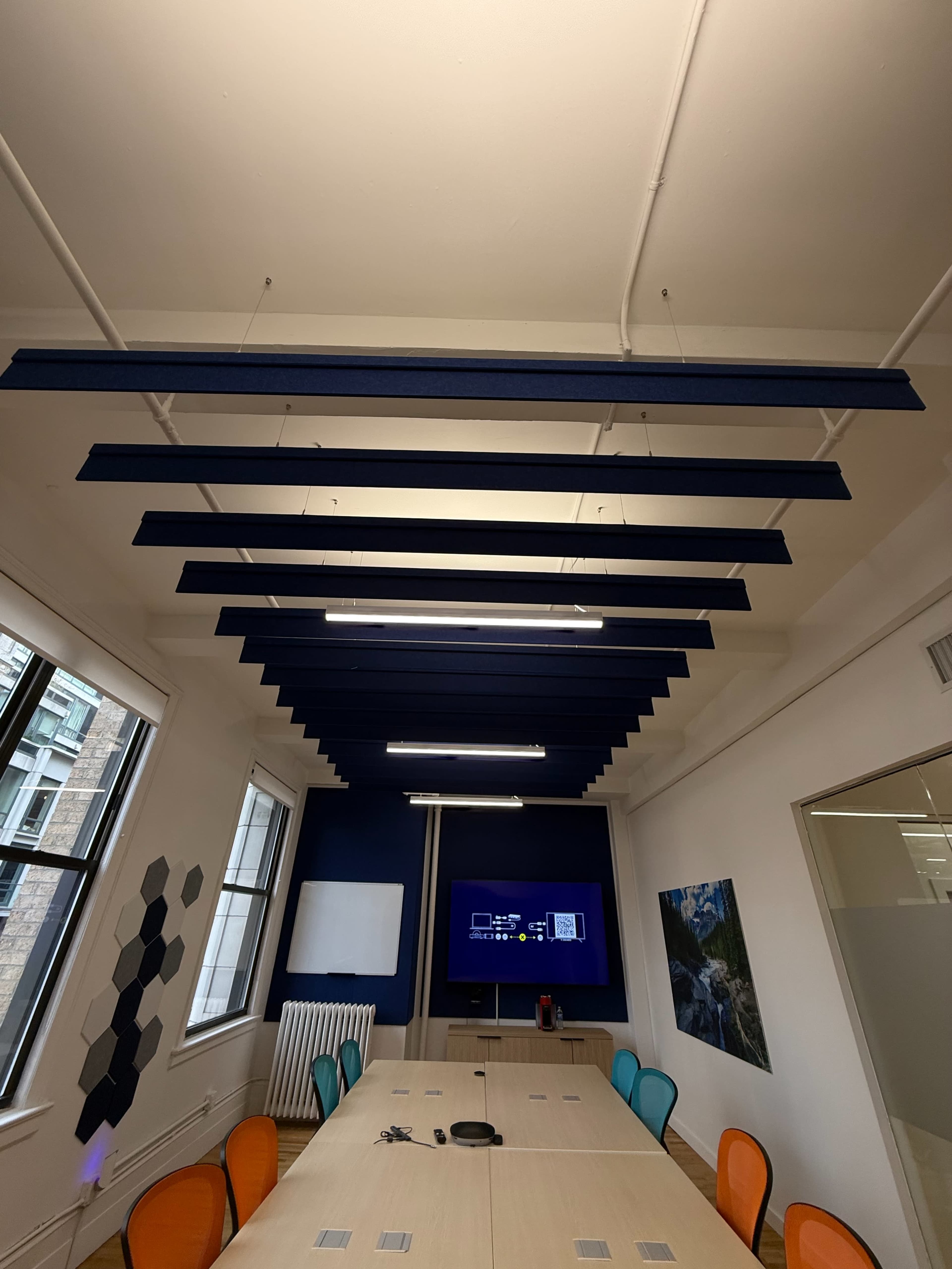 The image shows a modern conference room with a long table surrounded by colorful chairs and acoustic panels hanging from the ceiling.