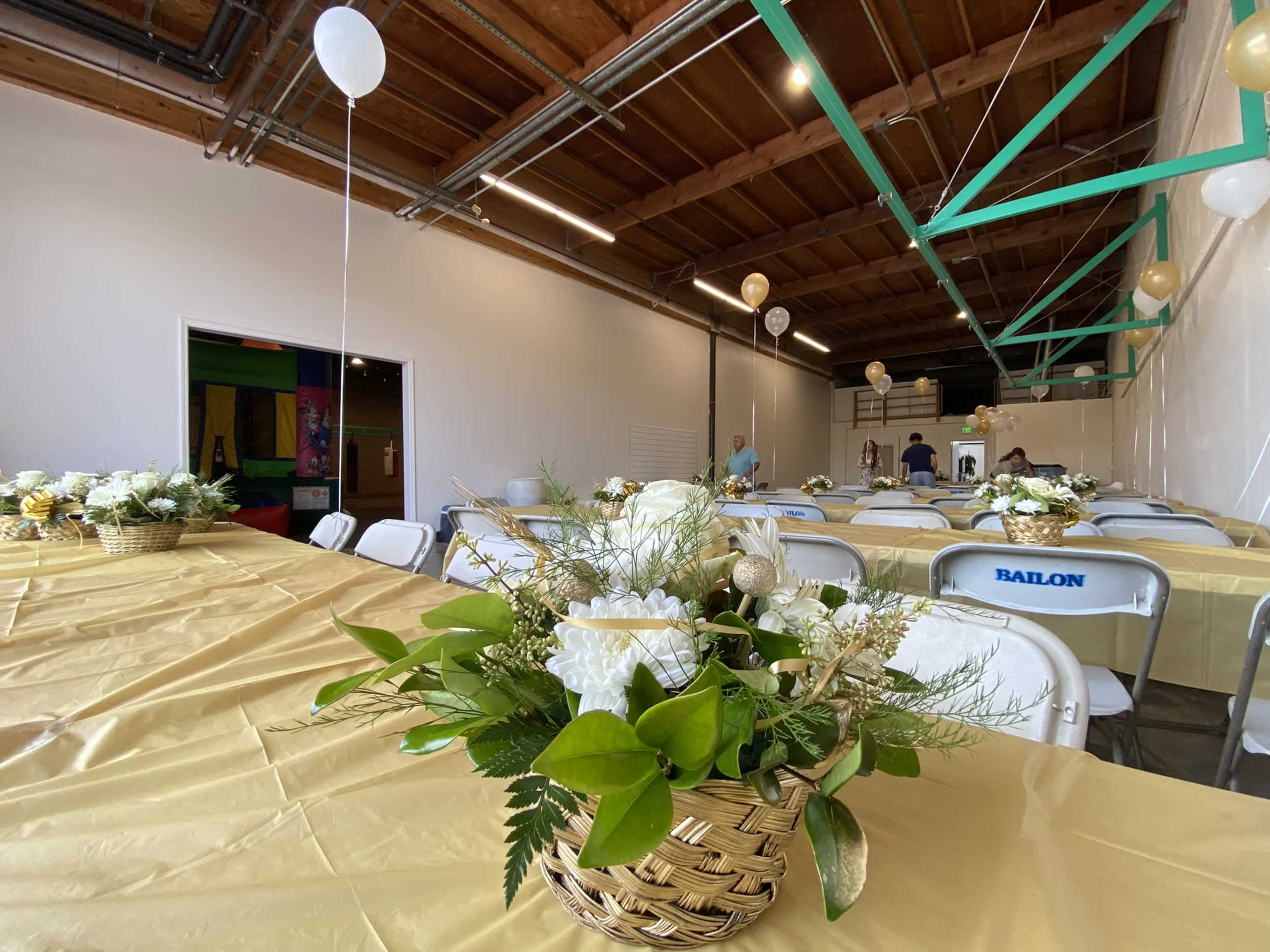 A spacious room with tables set for an event, adorned with floral centerpieces and balloons hanging from the ceiling.