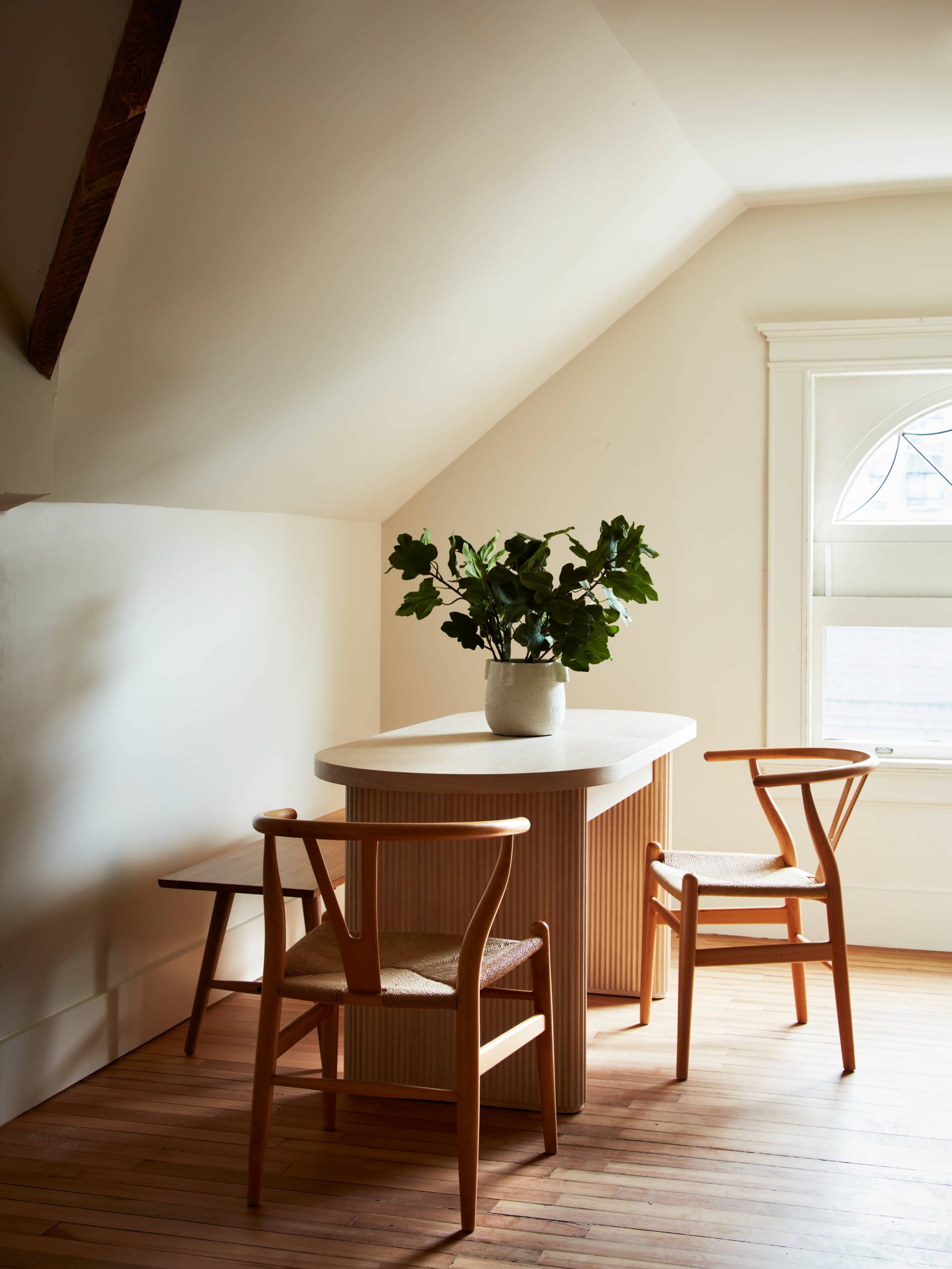 A small dining area features a round table with a textured surface surrounded by two wooden chairs and a bench, complemented by a vase of greenery.