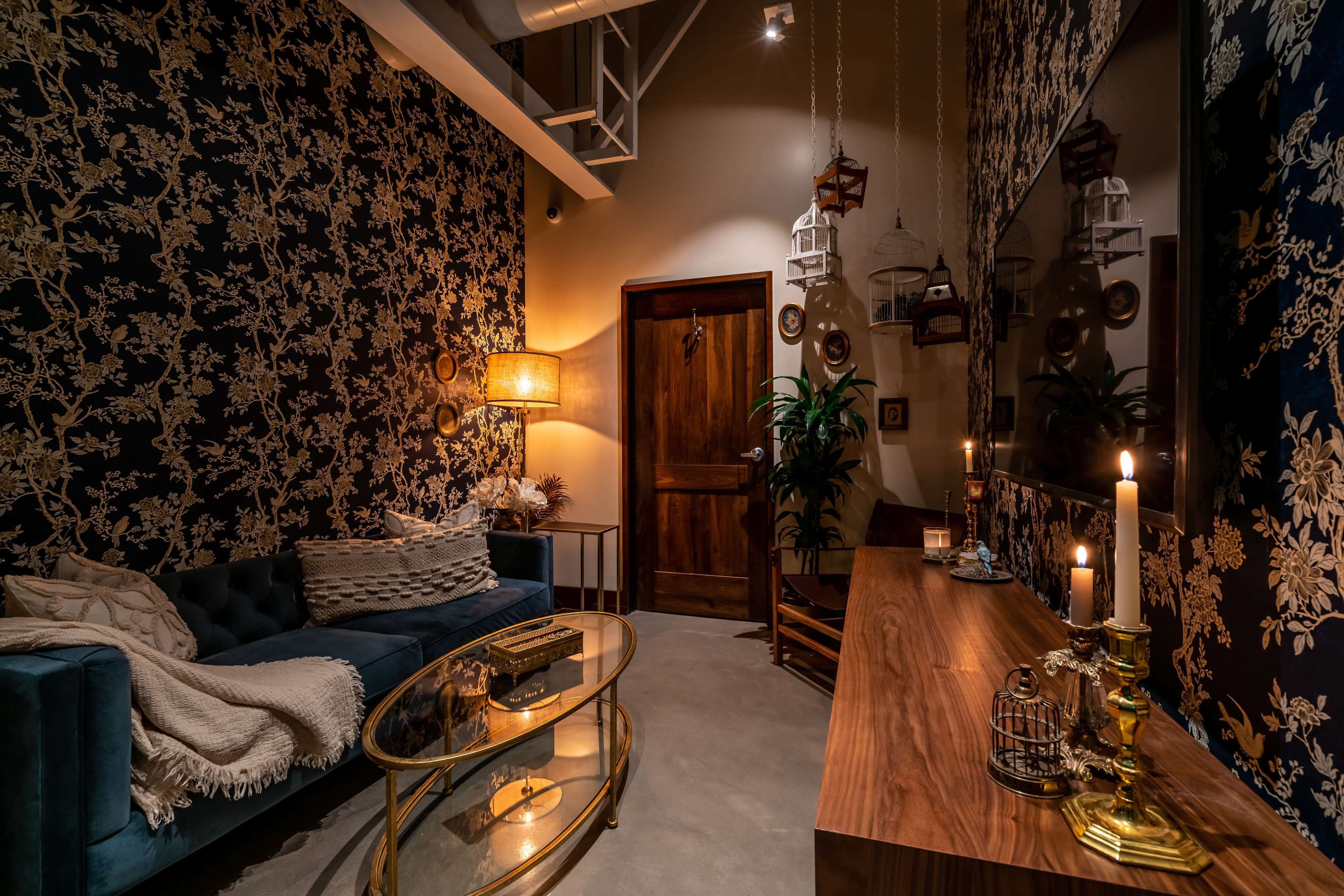 The image shows a cozy room with floral-patterned wallpaper, a blue velvet sofa, a wooden door, and a stylish wooden table adorned with decorative items and candles.