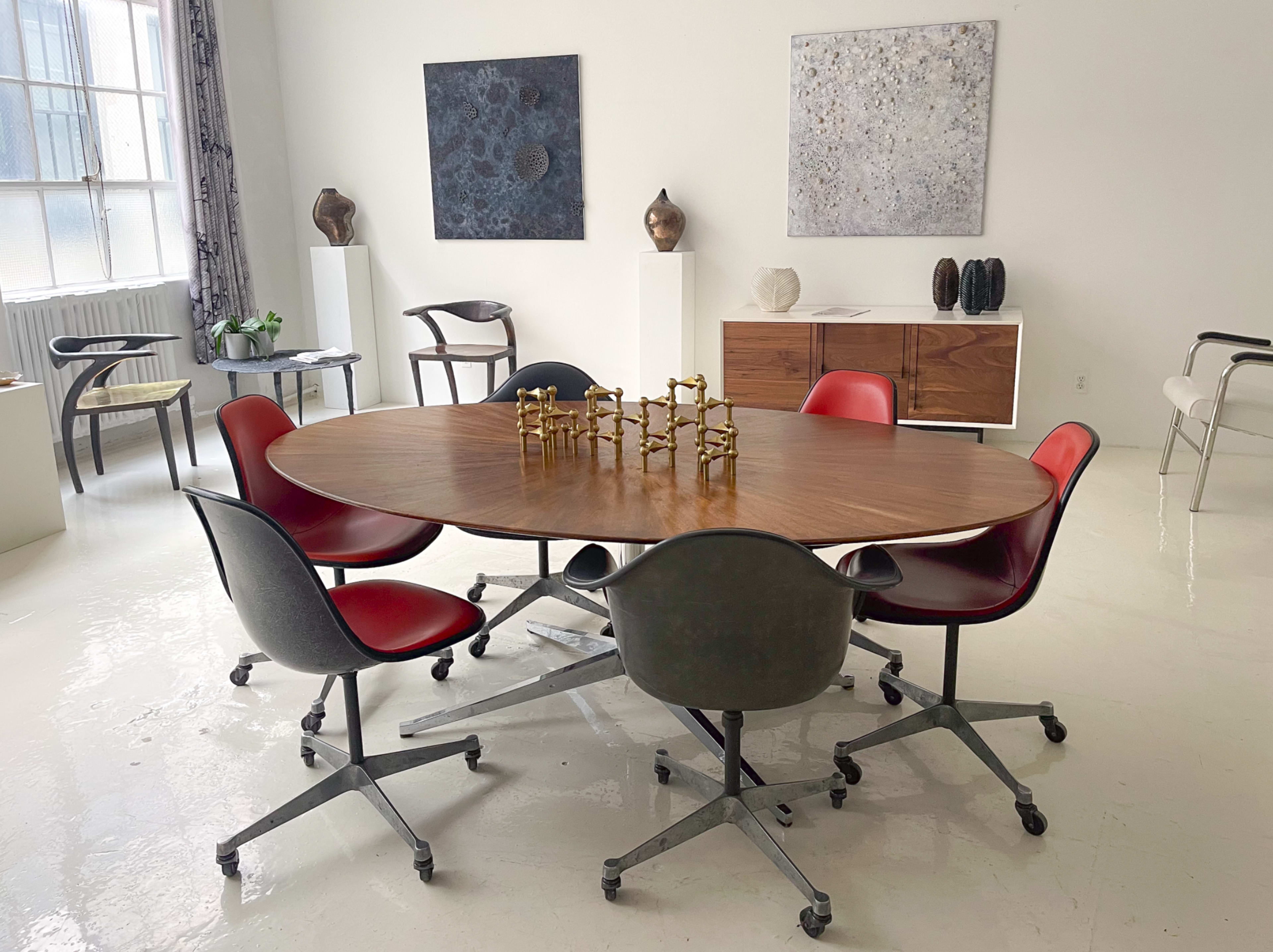 A modern conference room features a large oval wooden table surrounded by six chairs in various colors, with abstract artworks on the walls and decorative objects displayed on a sideboard.