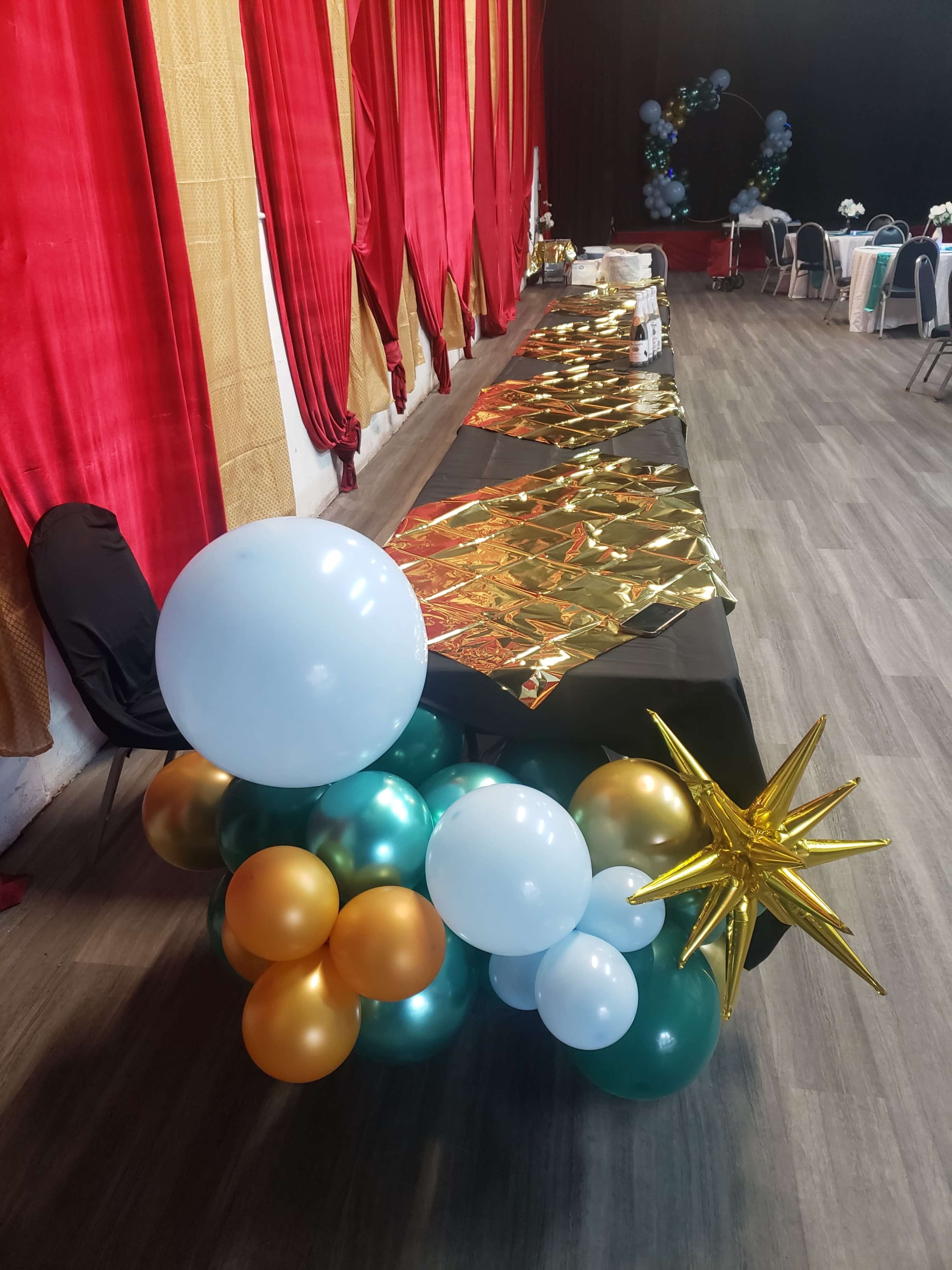 A table adorned with gold table covers and colorful balloon decorations is set in a venue with red curtains.