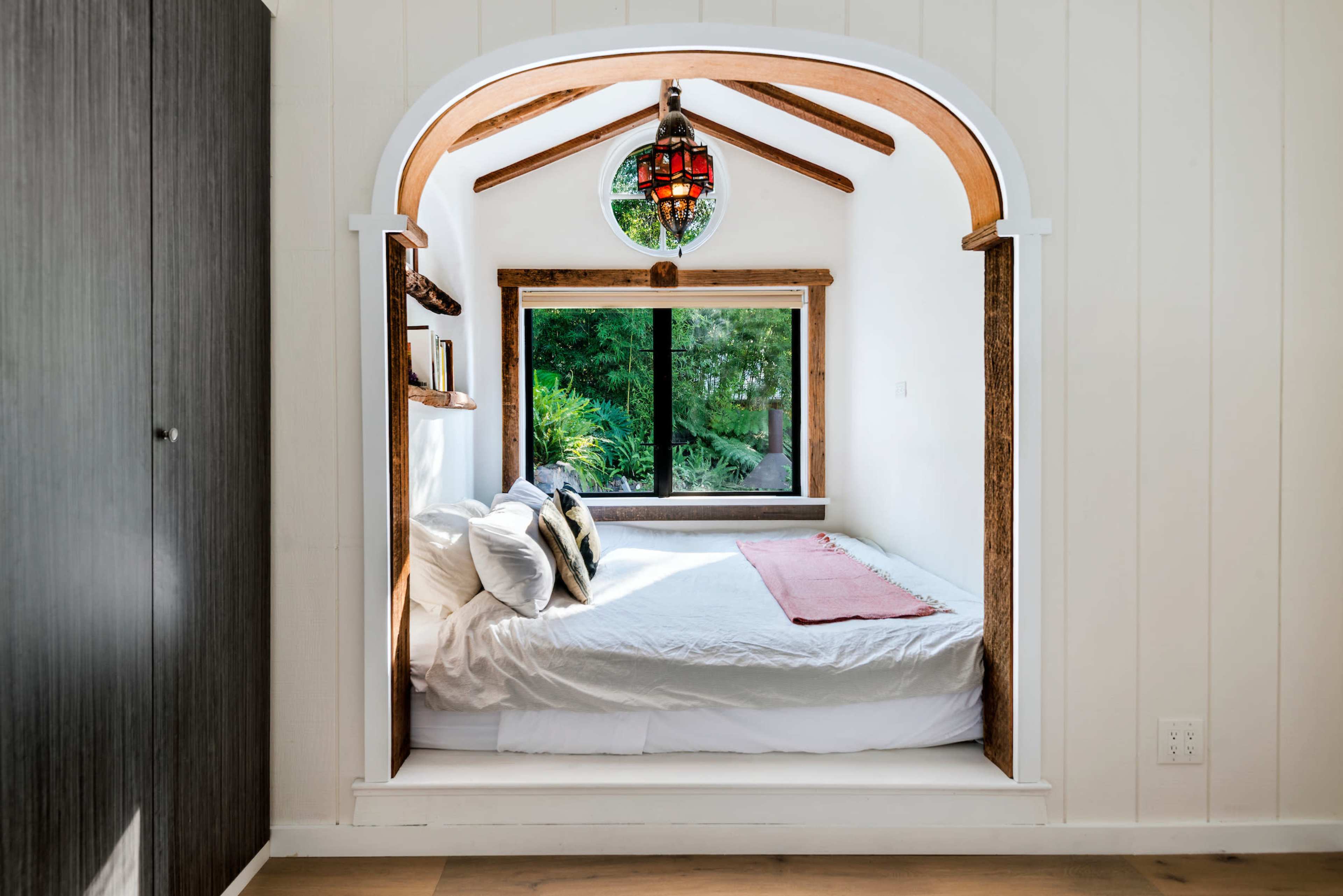 A small, cozy nook with a bed framed by white walls and wooden beams, featuring a large window that showcases greenery outside.
