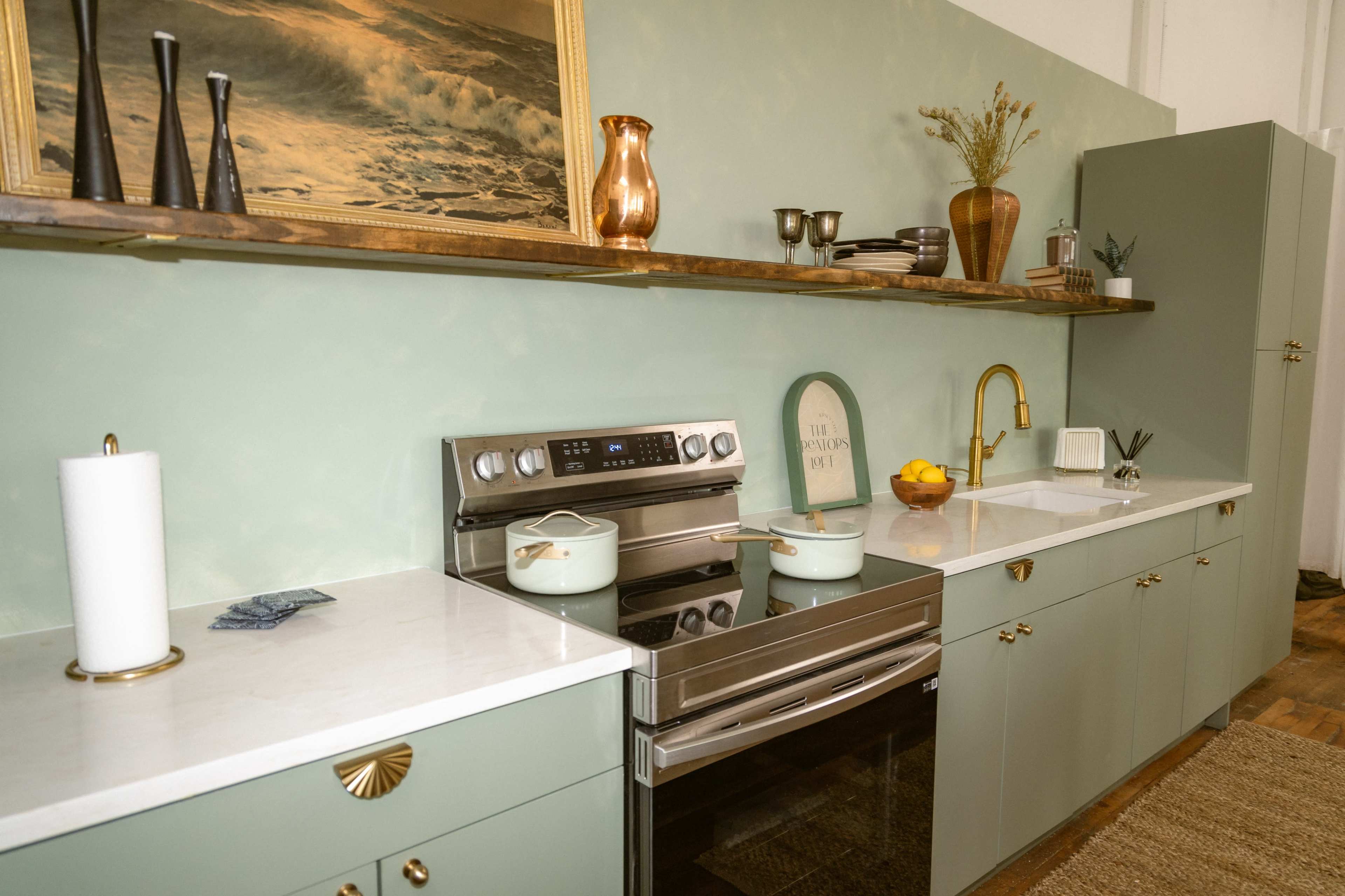The image shows a modern kitchen with green cabinets, a marble countertop, and stainless steel appliances, featuring a wooden shelf with decorative items above.