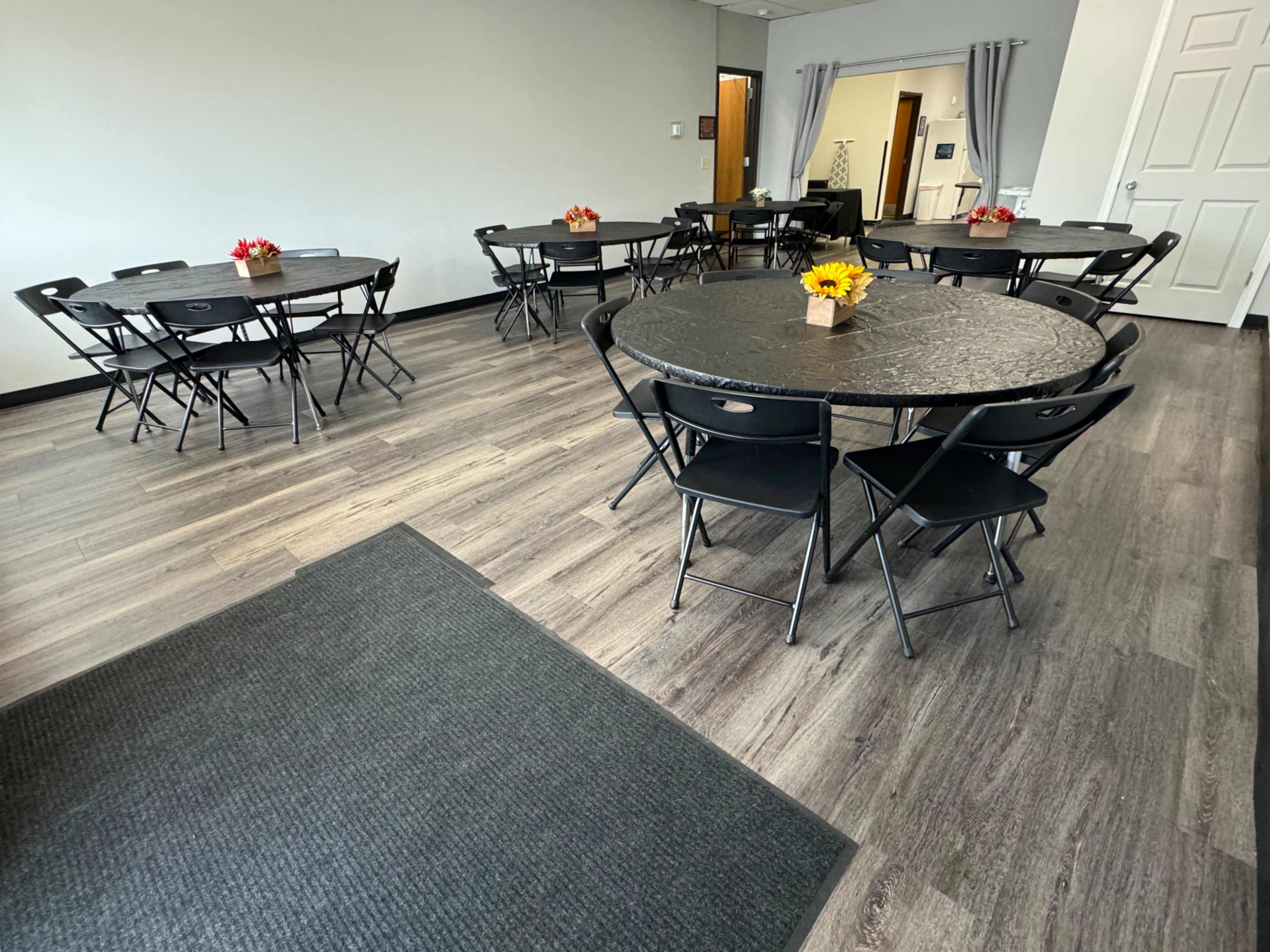 A well-lit meeting room with several round black tables set with small flower centerpieces, arranged on a wooden floor.