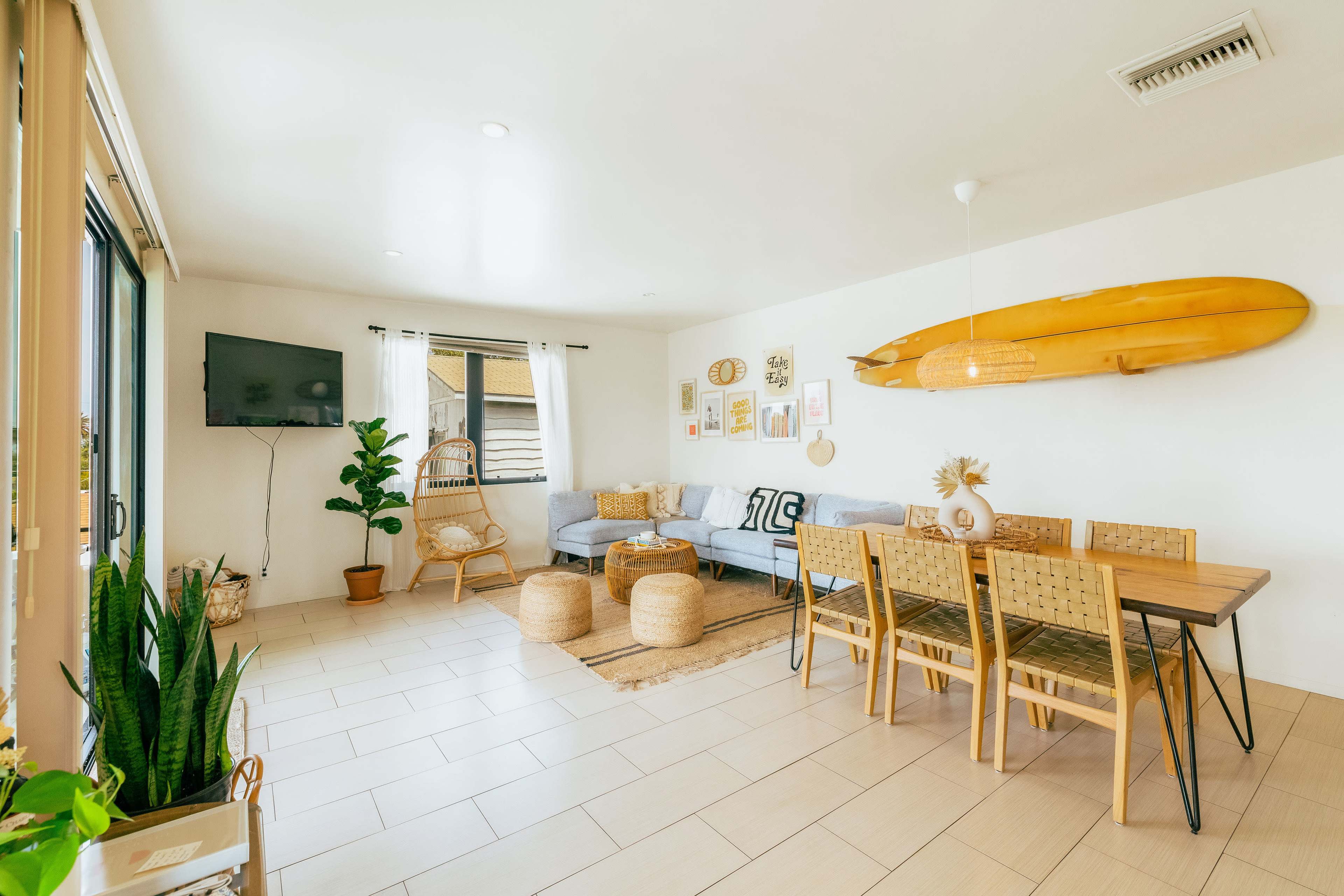 A bright, modern living space featuring a light blue sofa, a wooden dining table with chairs, and surfboard decor on the wall.