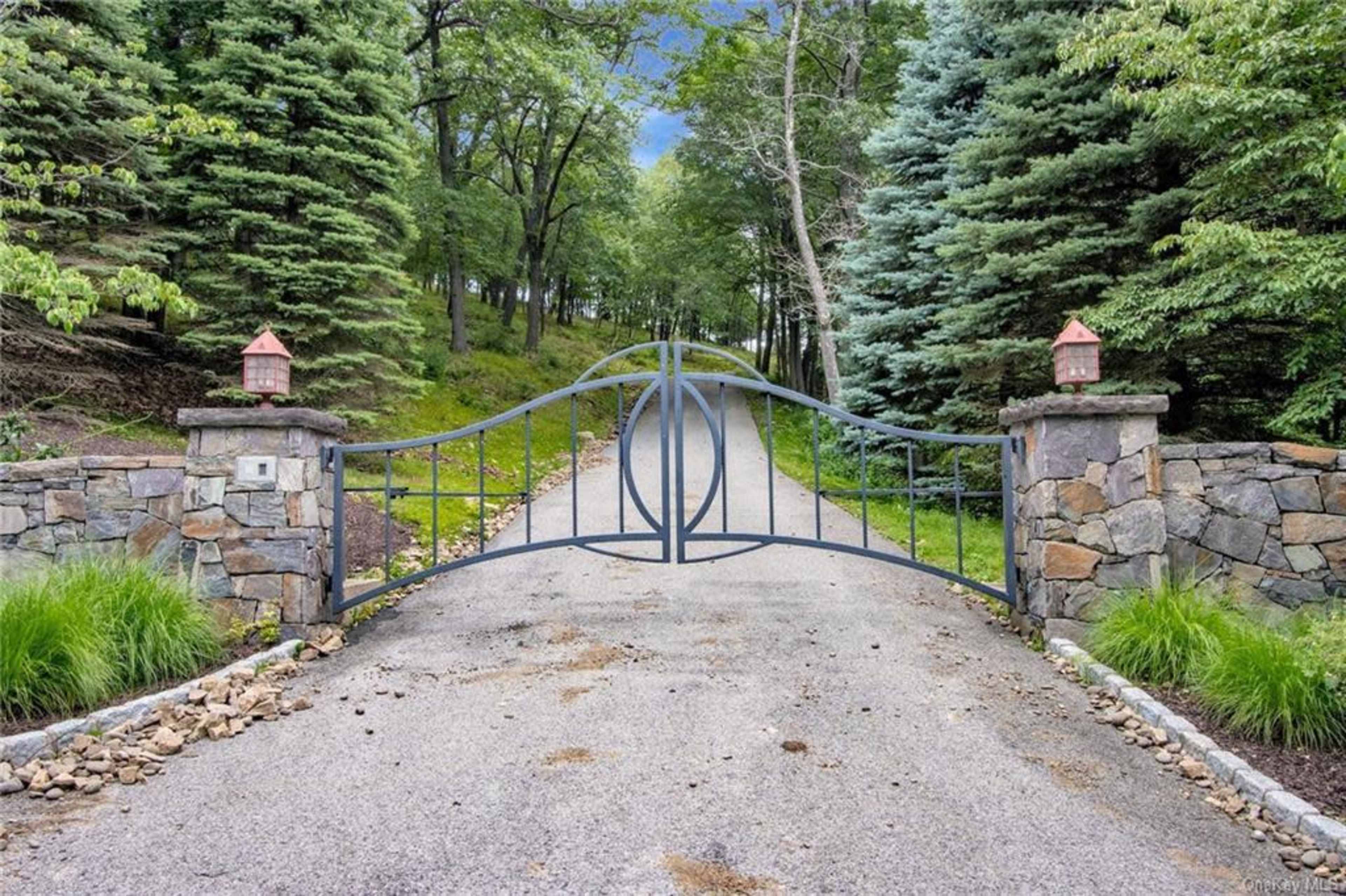 A large wrought iron gate stands at the entrance of a gravel driveway, flanked by stone pillars and surrounded by trees.