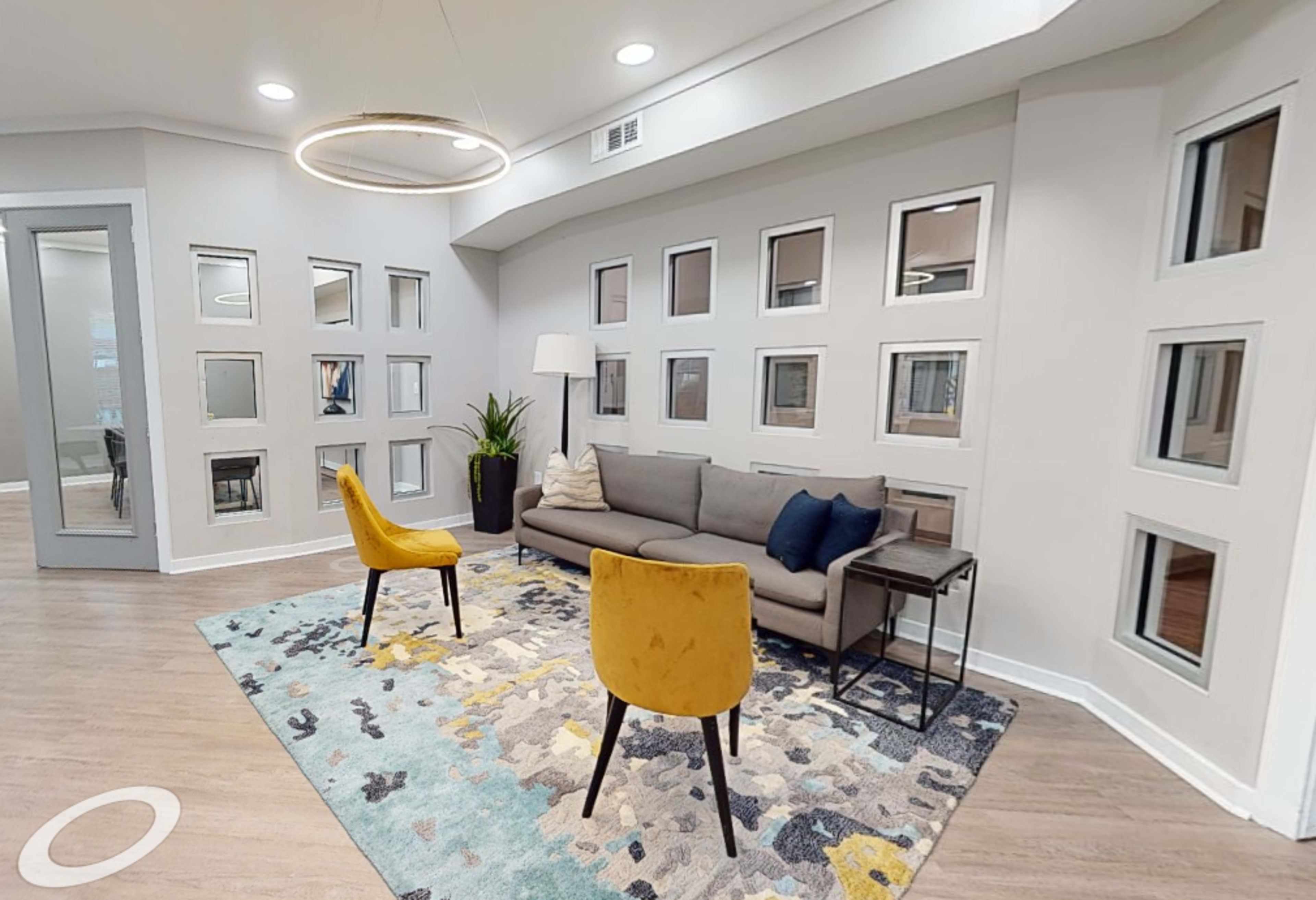 The image shows a modern lounge area featuring a gray sofa, two yellow chairs, a small side table, and a colorful area rug against a wall with multiple square windows.