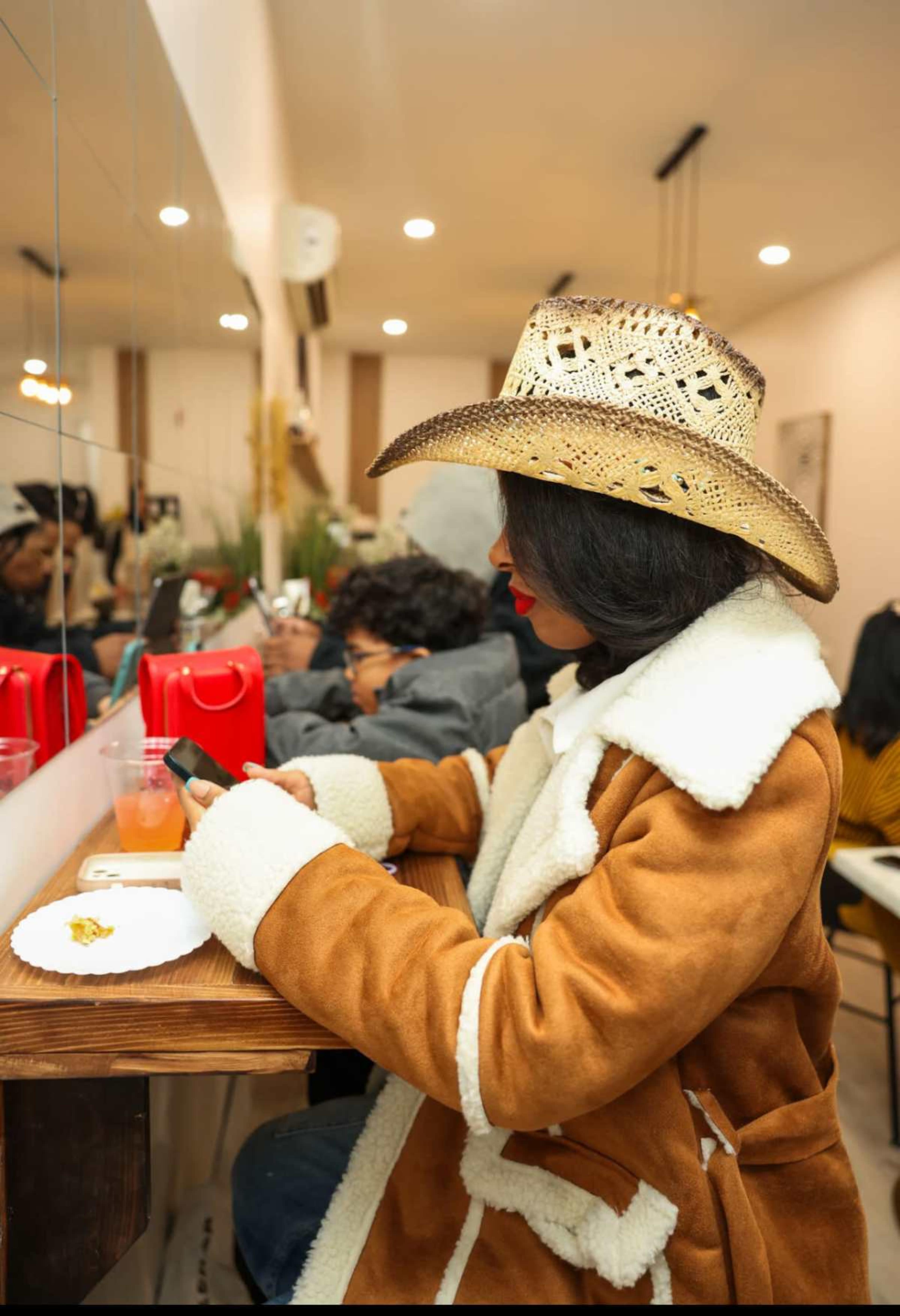 A woman wearing a cowboy hat and a brown coat sits at a counter, looking at her phone while a person in the background enjoys a drink.