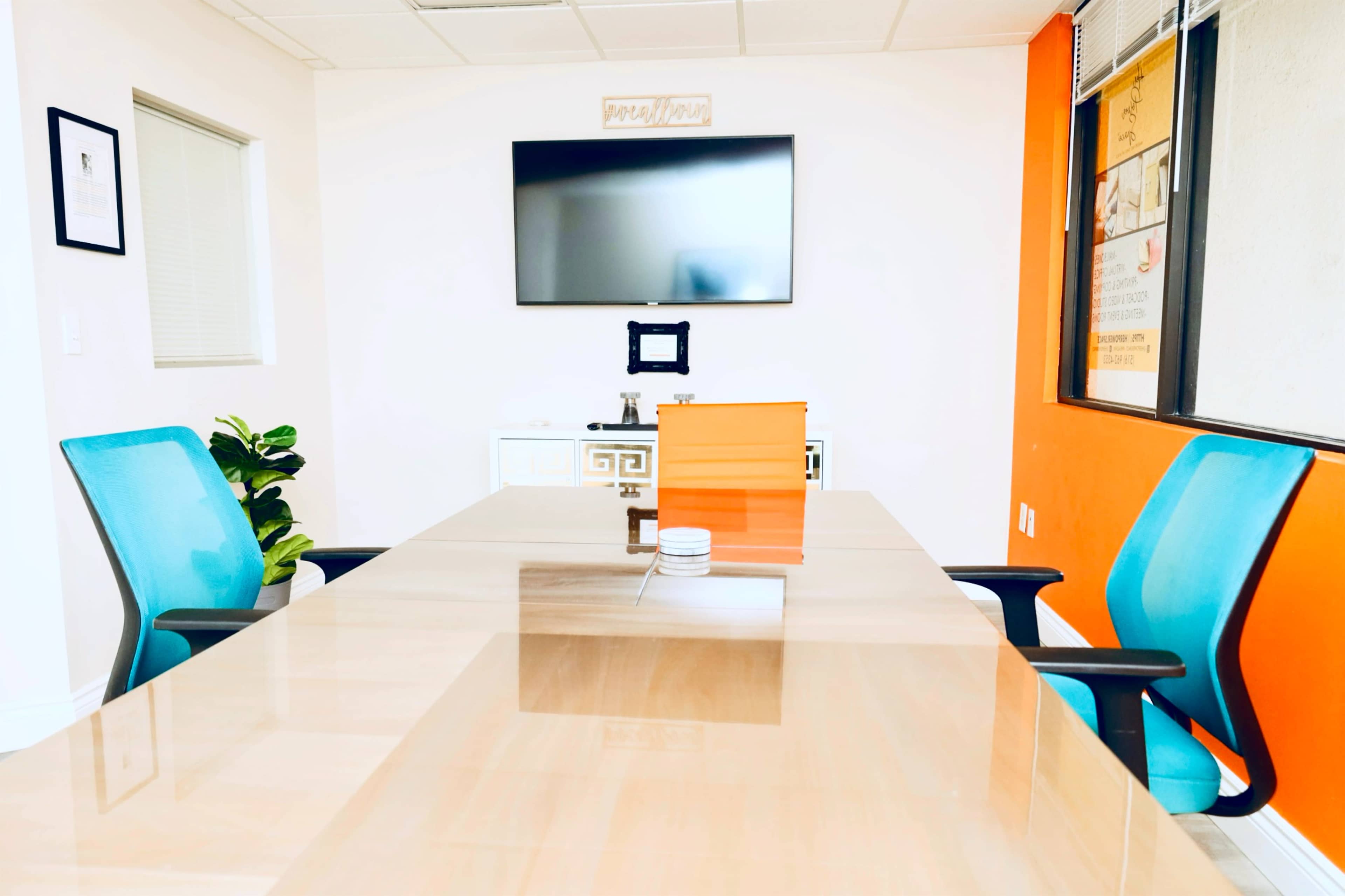 A modern conference room features a glass table with two blue chairs, a flat-screen TV on the wall, and an orange accent wall.