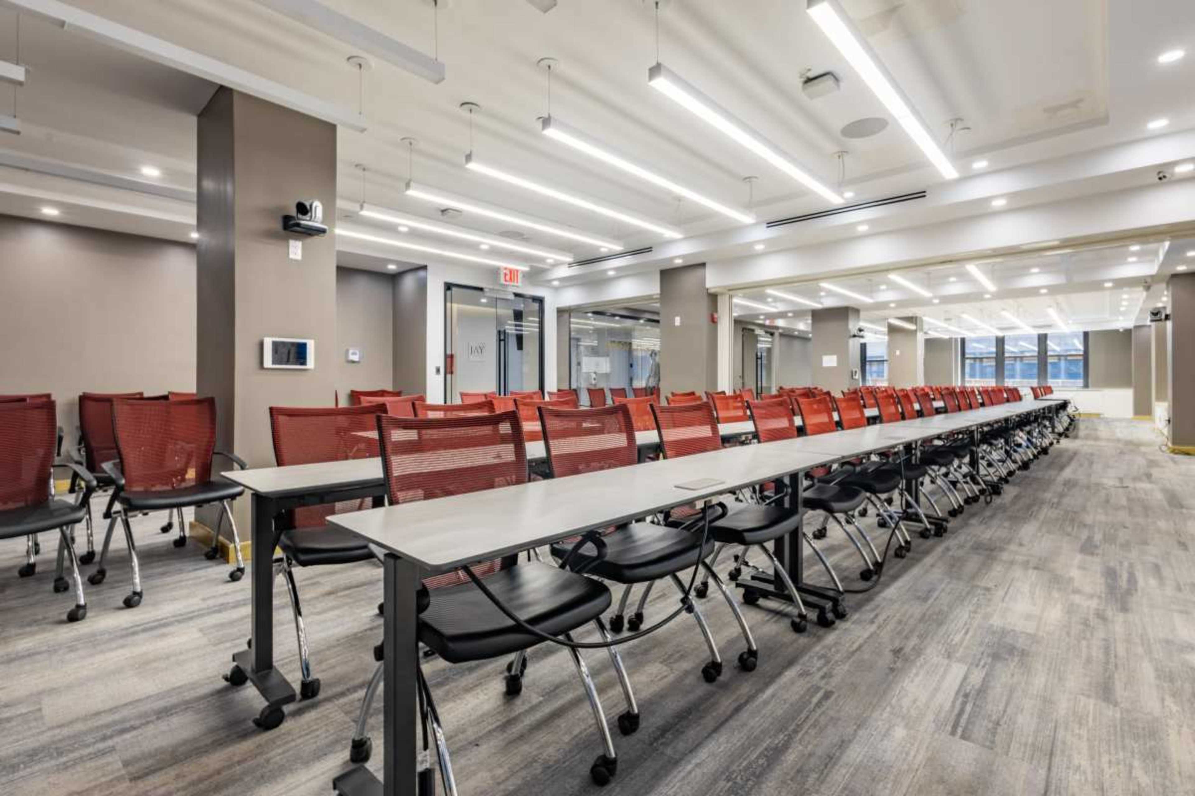 The image shows a modern conference room arranged with rows of red mesh chairs and tables, illuminated by overhead lights.