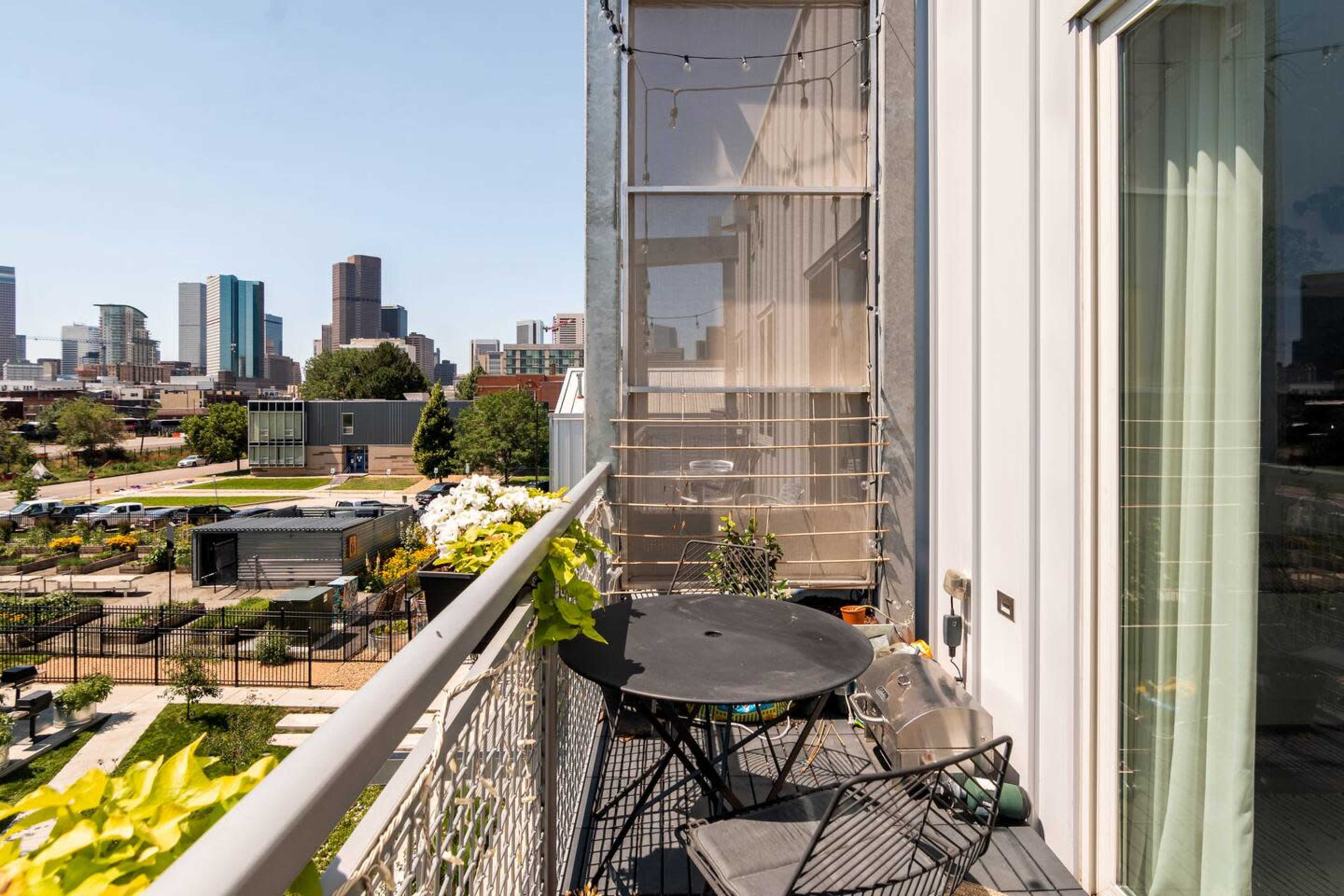 A small balcony features a round black table and chairs, overlooking a cityscape with buildings and greenery below.