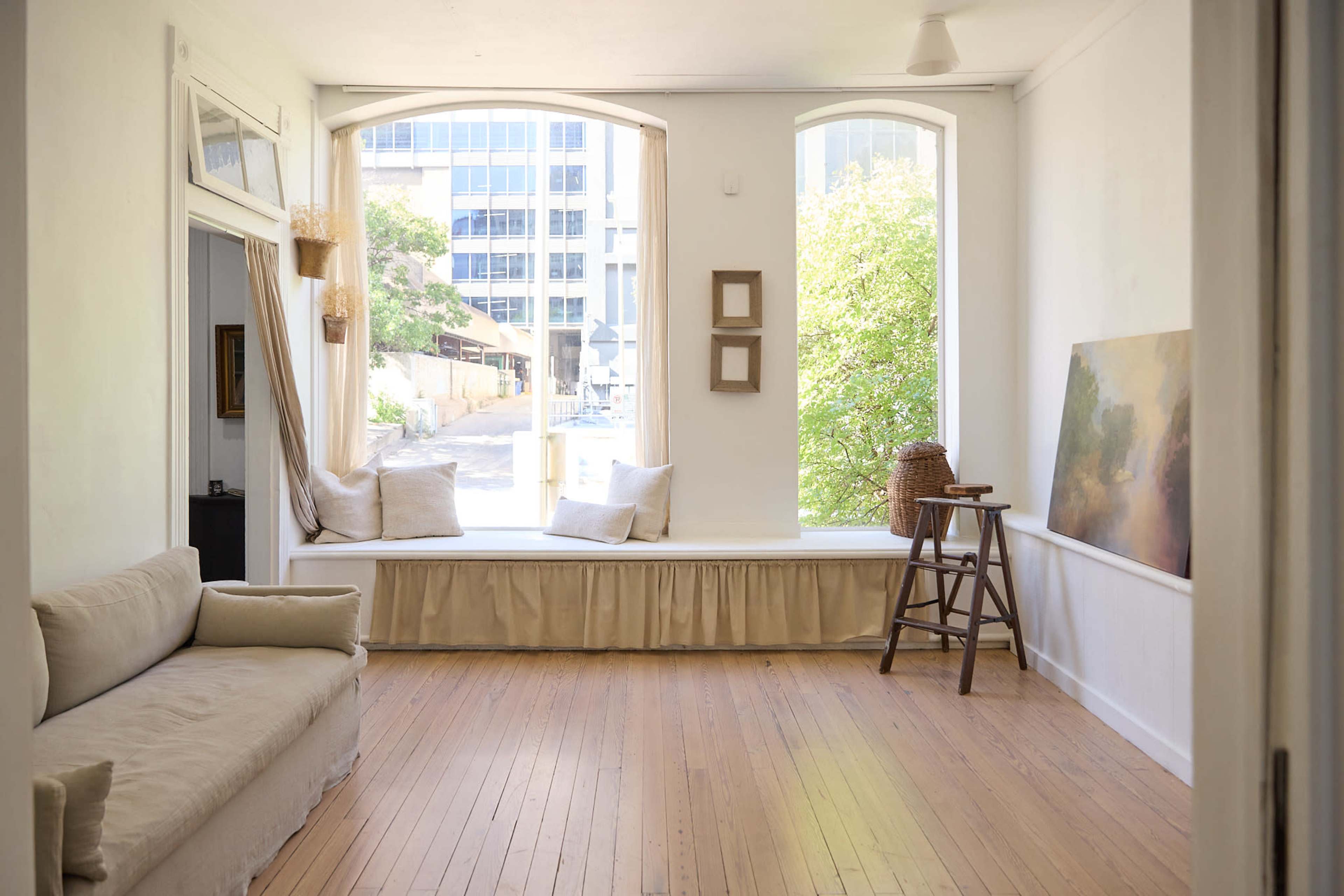The image shows a bright, minimalist room with large windows, a beige couch, and a small wooden stool beside a window seat adorned with cushions.