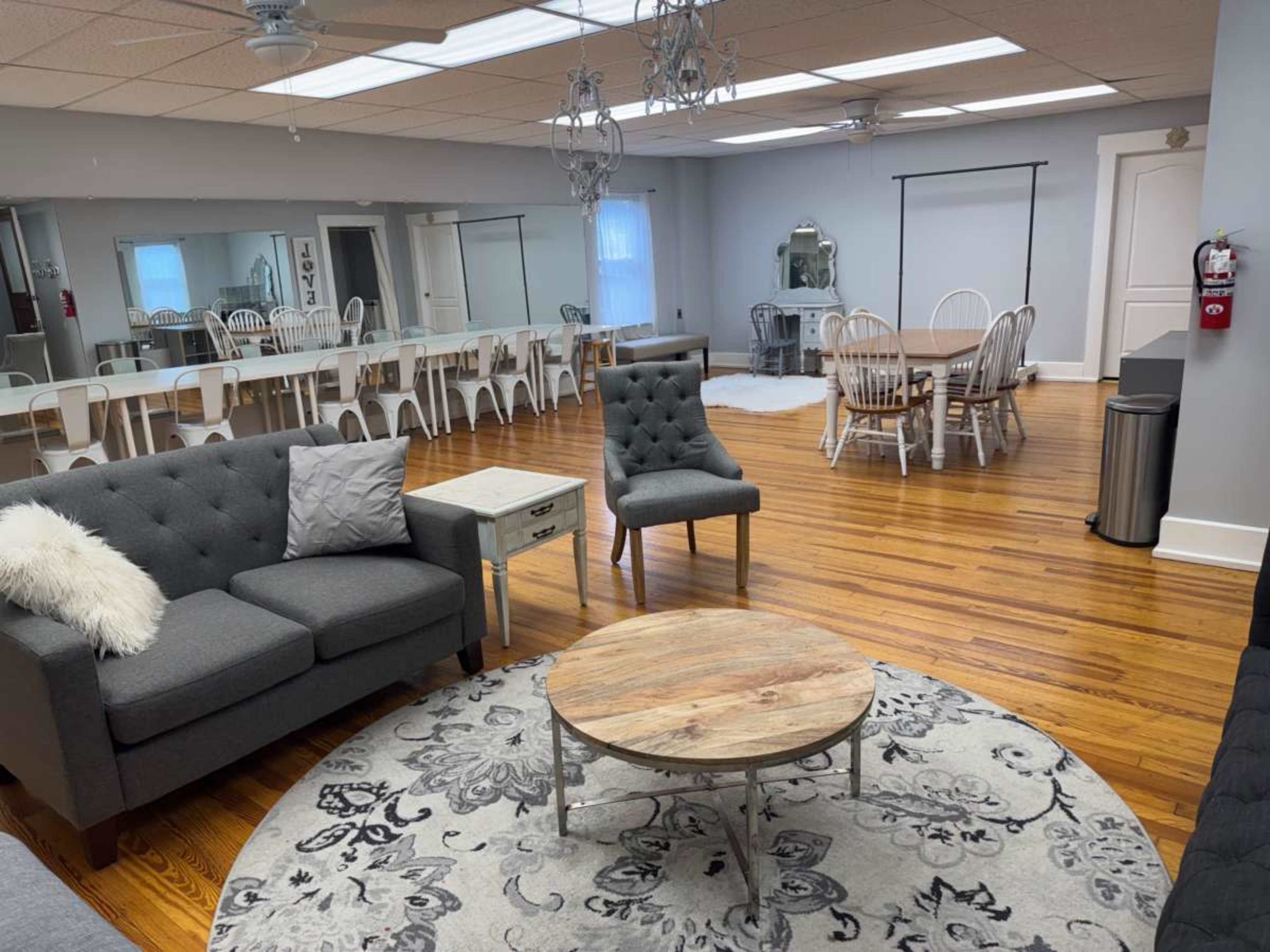 The image shows a spacious room with seating arrangements including a sofa and chairs, a round wooden coffee table, and a dining area with tables and chairs in the background.