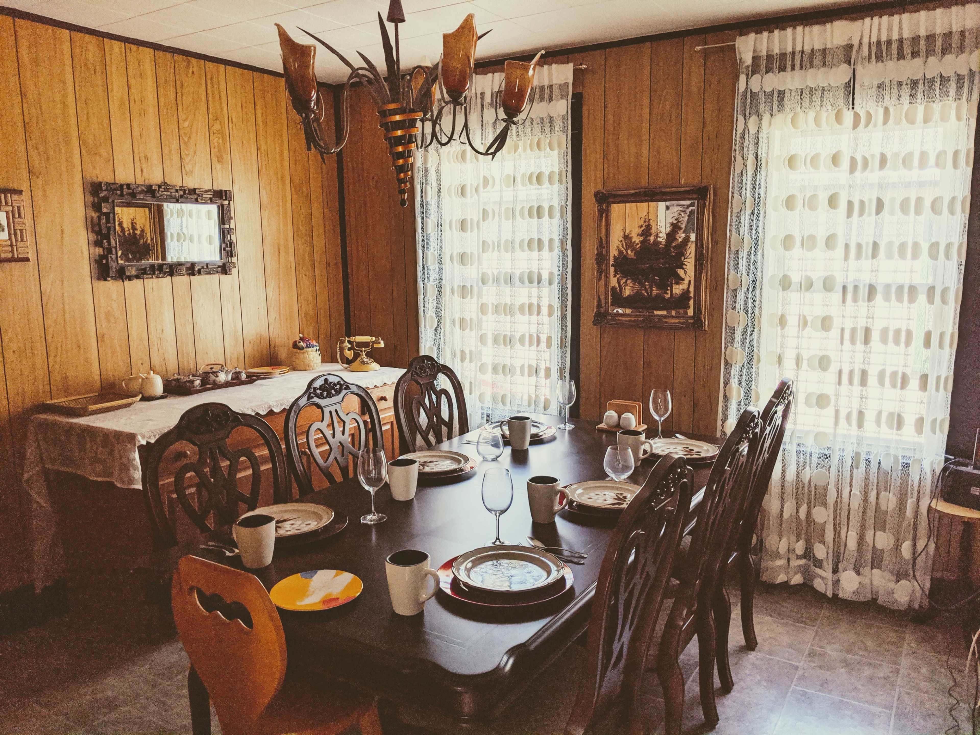 A wooden dining room features a large table set for a meal, surrounded by ornate chairs, with a chandelier overhead and windows dressed in polka-dotted curtains.