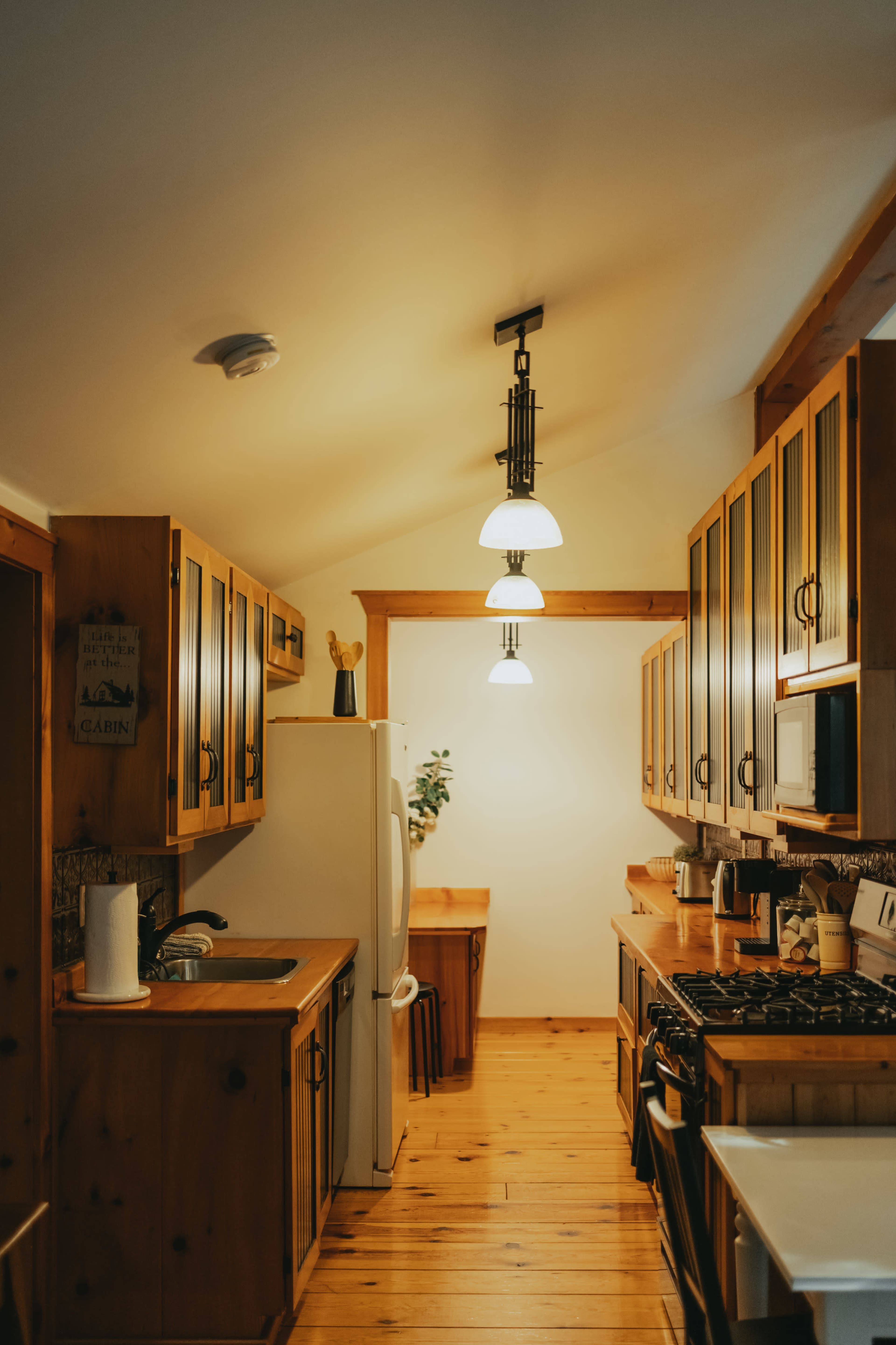 A wooden kitchen with cabinets, a white refrigerator, and a stove, illuminated by hanging light fixtures.