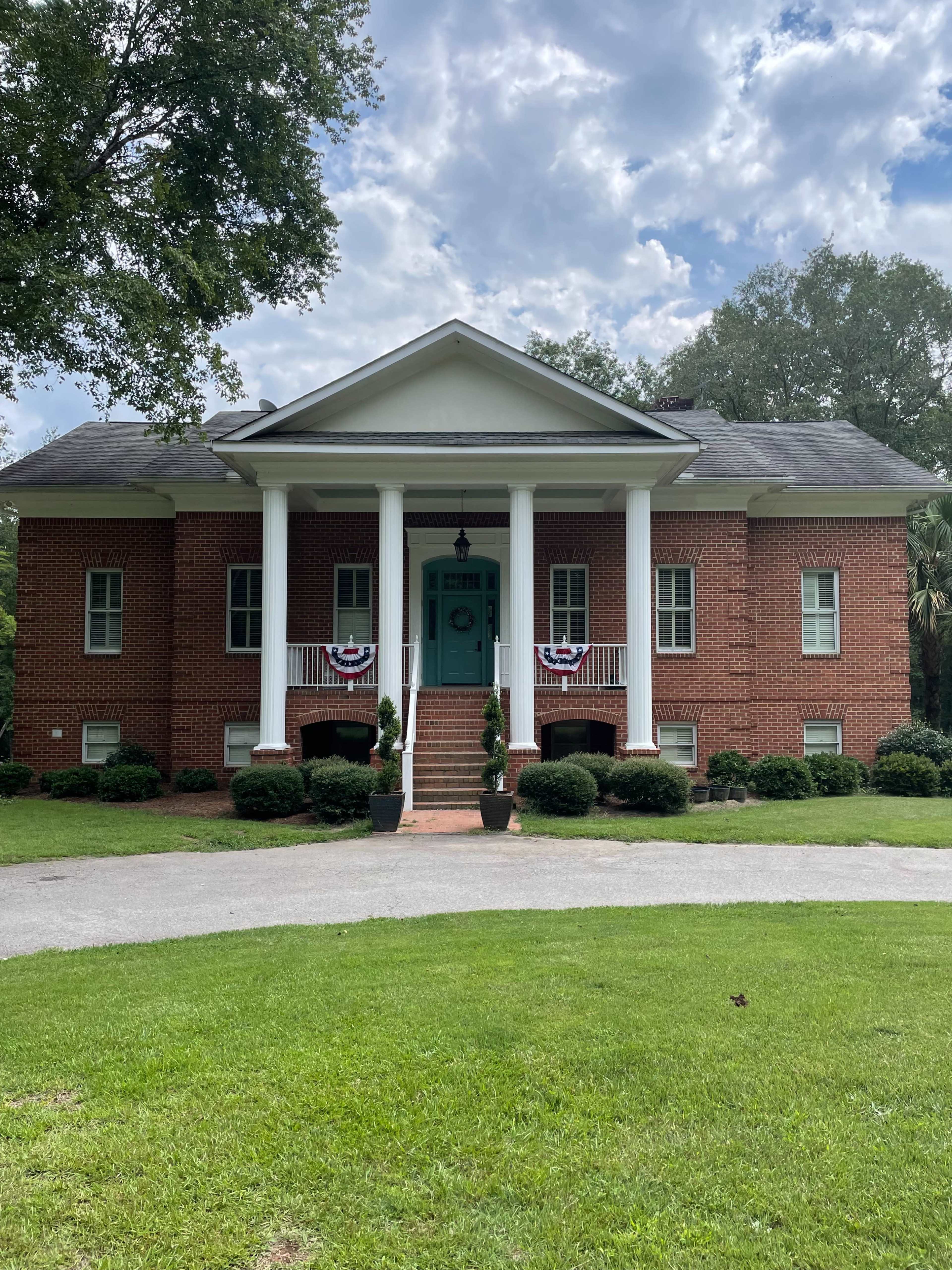 A two-story brick house with white columns, a green door, and decorative bunting on the front porch is set on a grassy lawn.