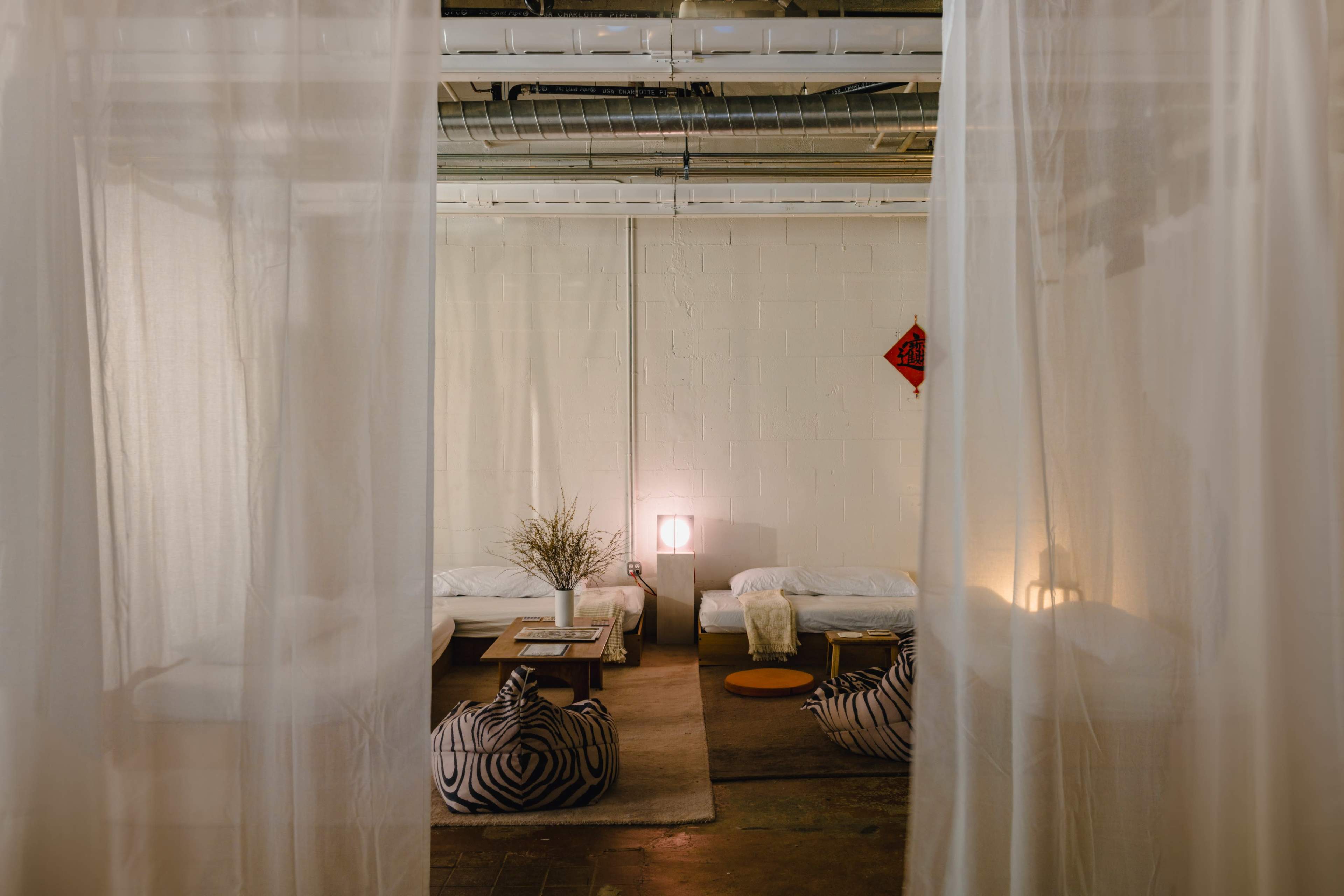 The image depicts a cozy, minimalist interior space with soft lighting, featuring sheer curtains, low seating, and decorative cushions.
