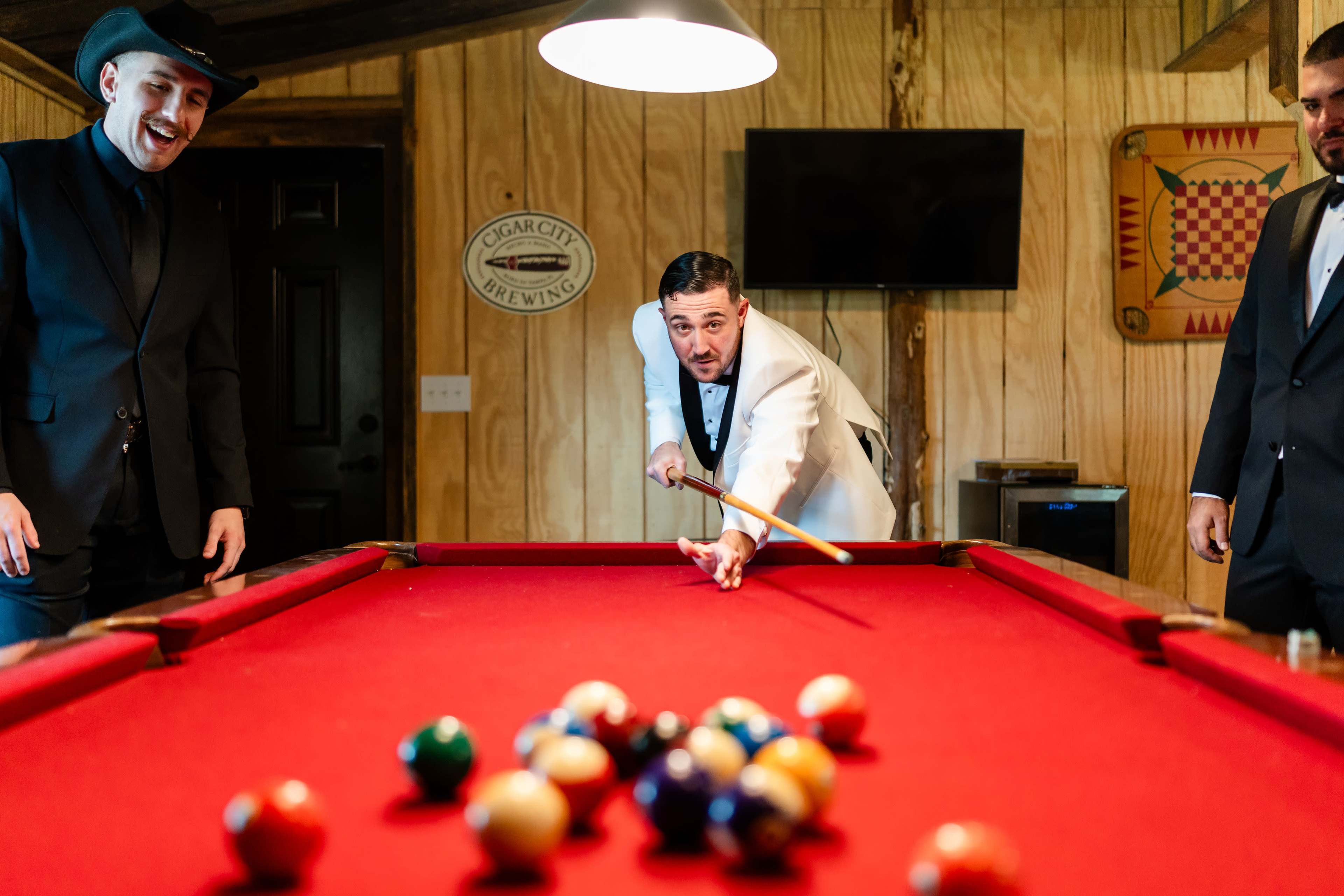 A man in a tuxedo prepares to take a shot at a pool table, while two other men look on in a wooden-paneled room.