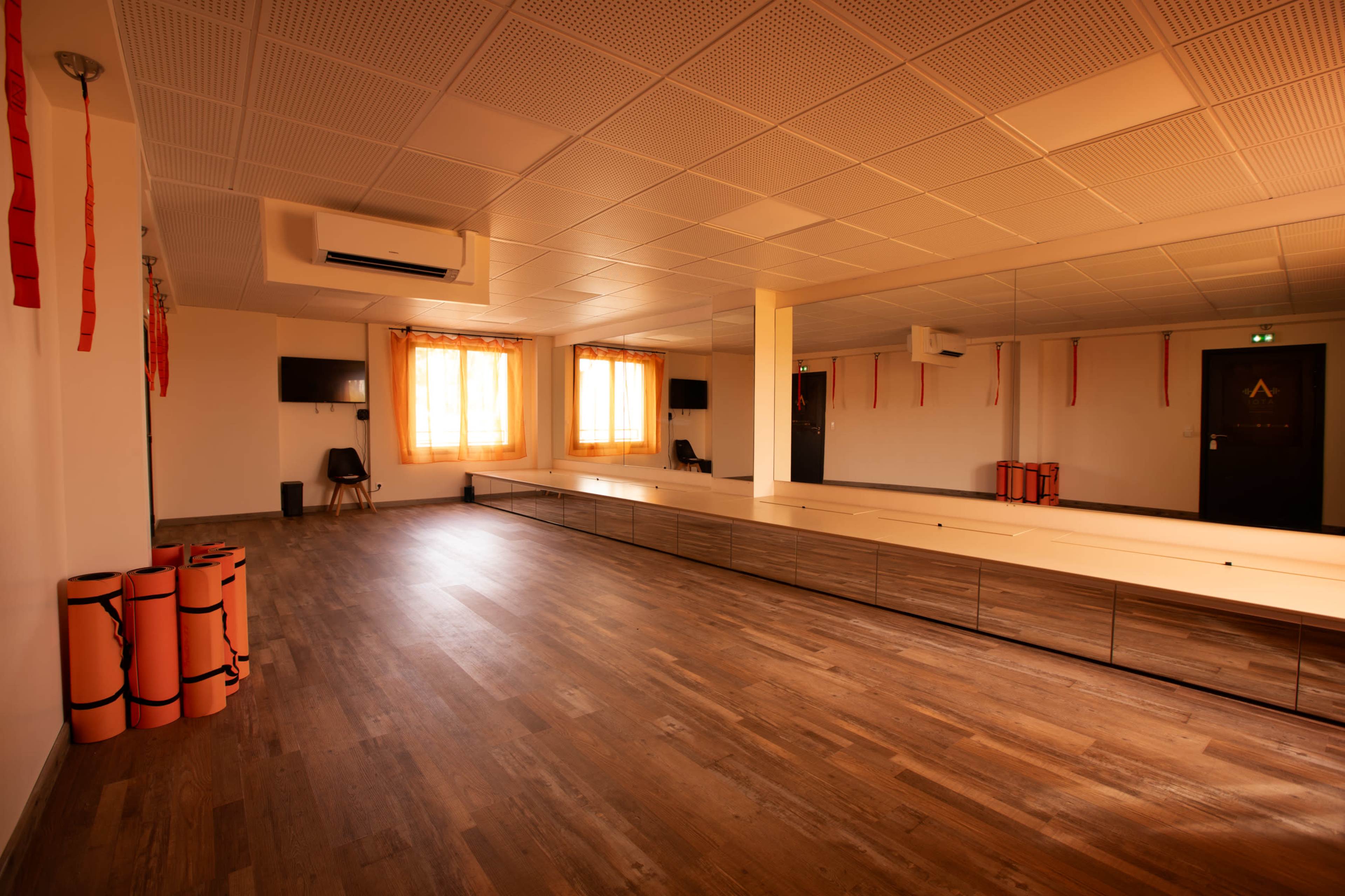 The image shows a spacious, well-lit room with wooden flooring, large mirrors along one wall, and exercise mats rolled up in orange.