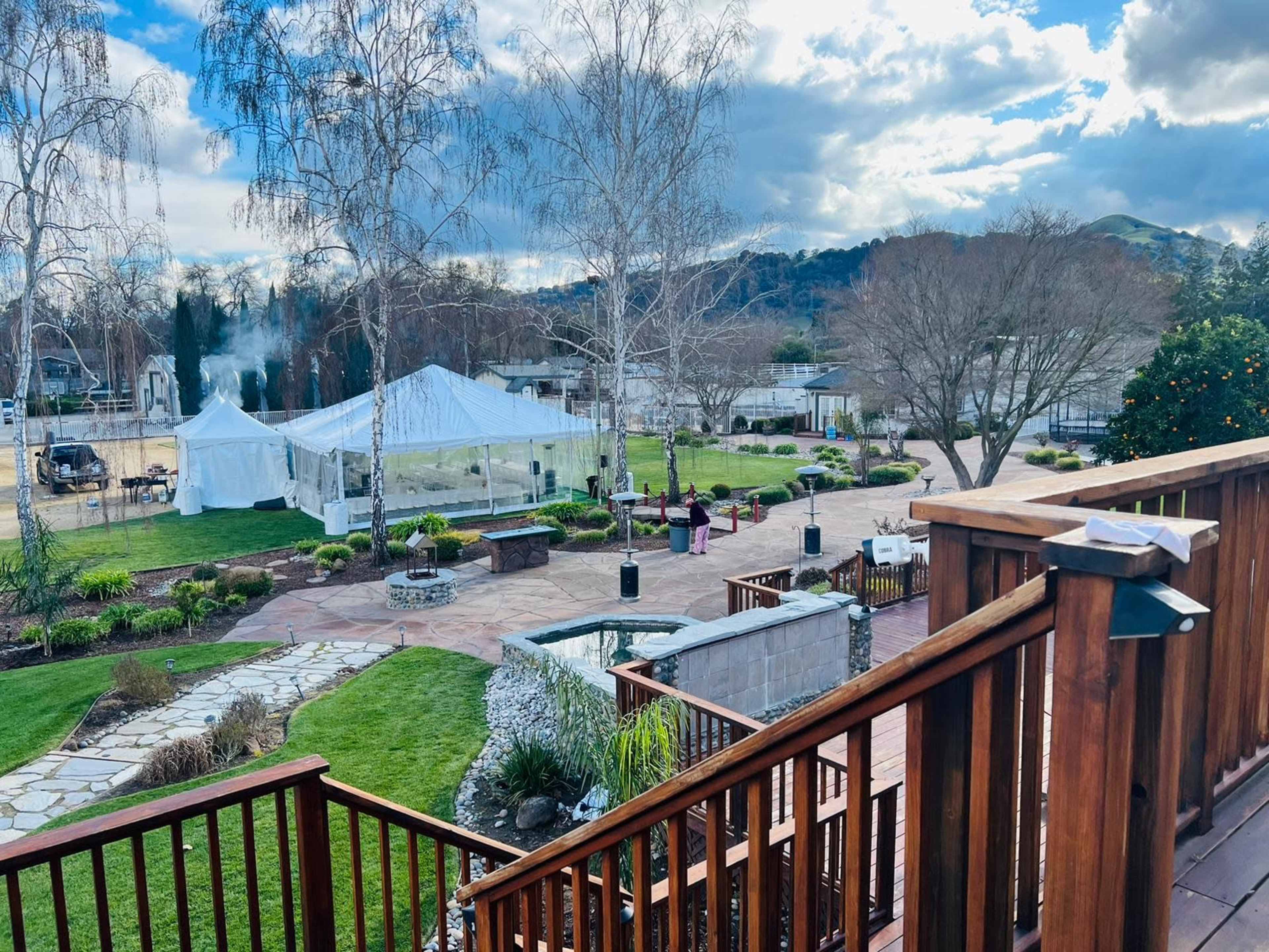 A spacious outdoor venue features a large white tent, landscaped gardens, and a pathway leading to a fountain, with mountains visible in the background.
